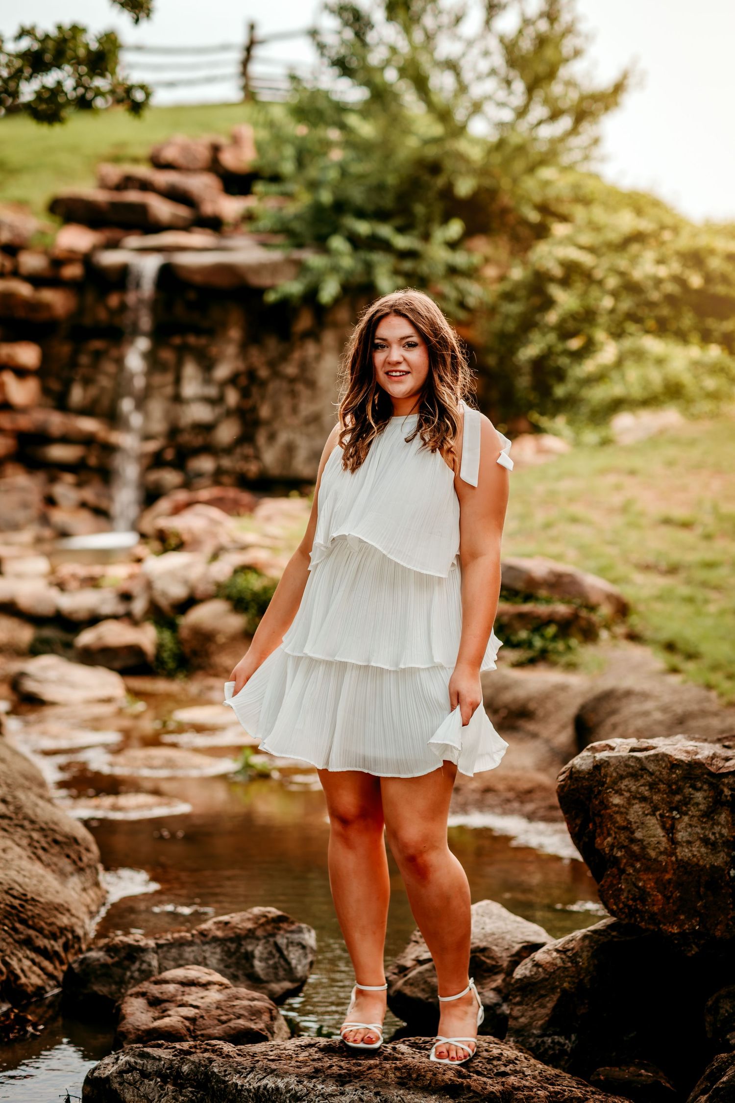 Young woman in white tiered dress standing on rocks near a small waterfall in a lush garden setting.