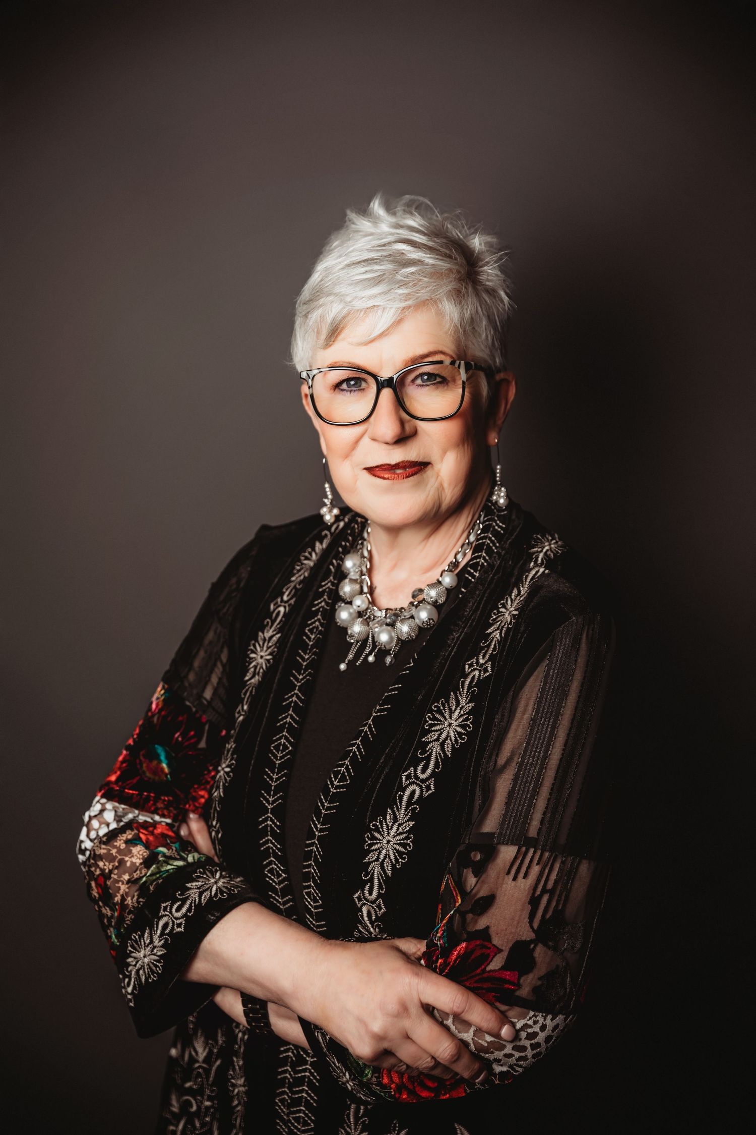 Elegant older woman with short gray hair, glasses, and floral black outfit poses confidently with arms crossed.