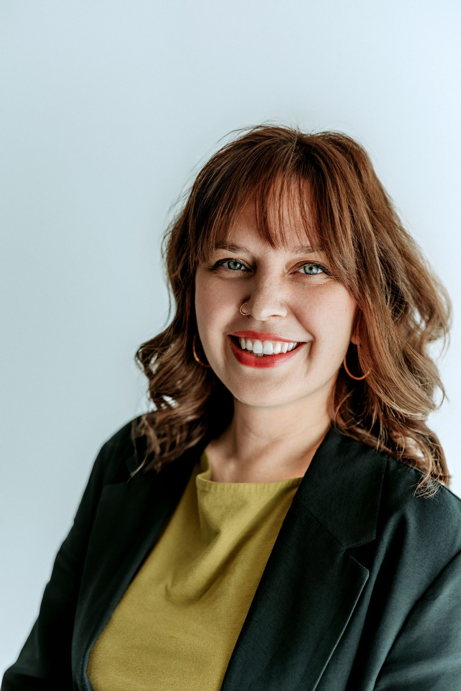 Smiling woman with auburn hair wearing a green blazer and yellow top against a light gray background.