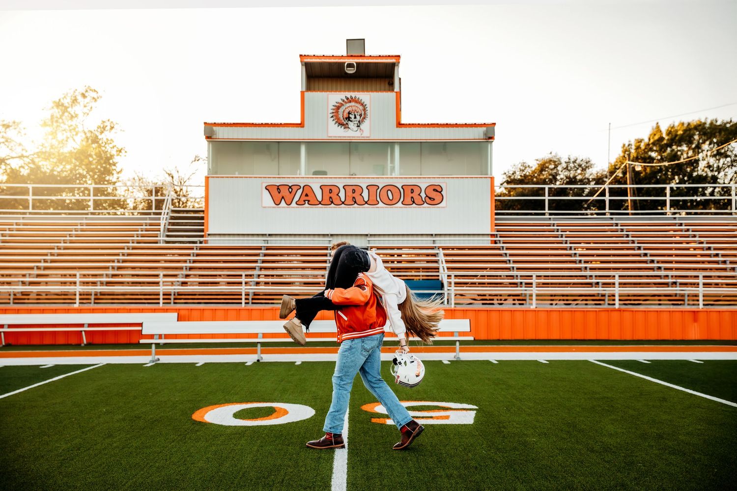 Couple embracing on Warriors football field at sunset, with orange stadium bleachers in the background.
