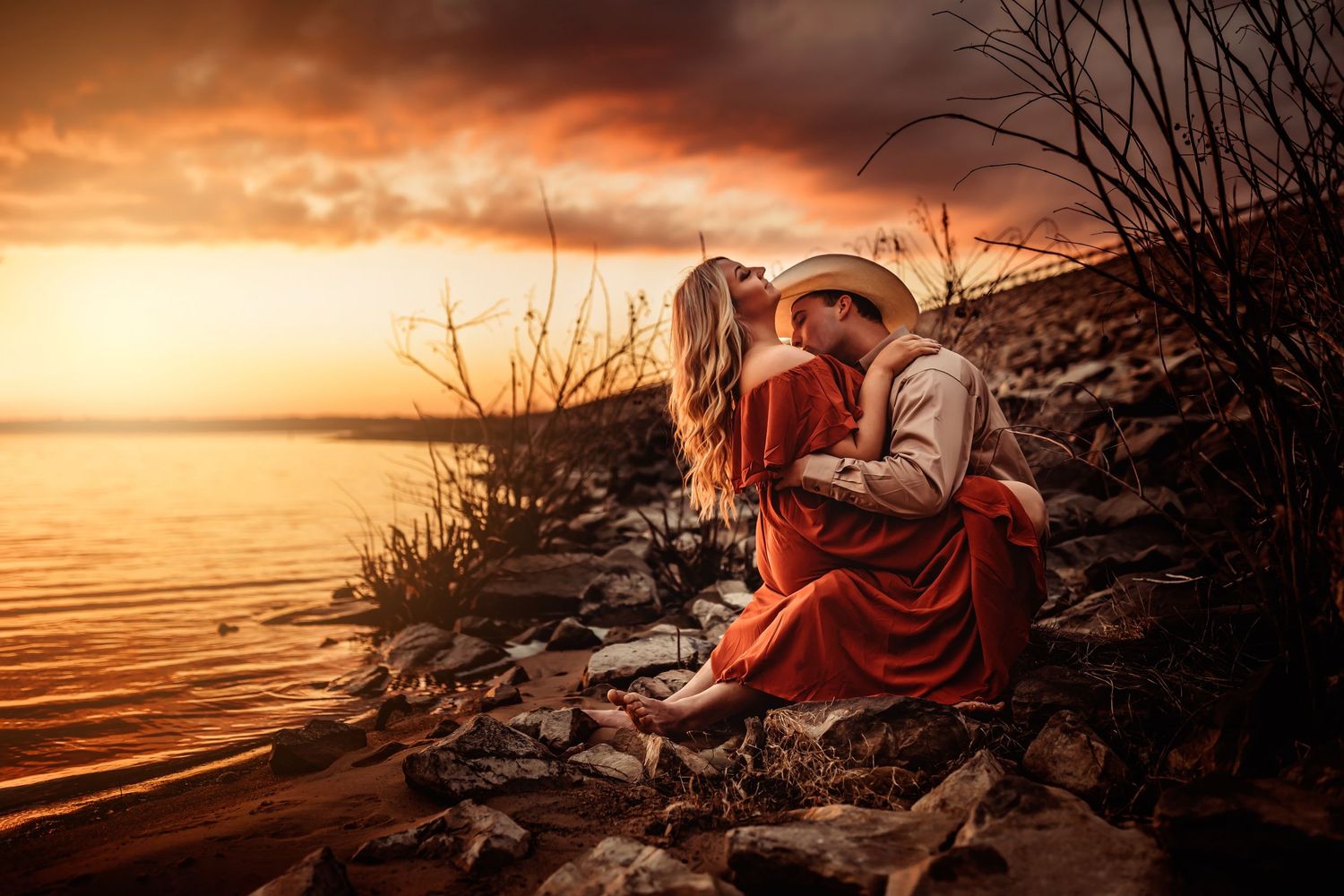 Couple sharing a romantic kiss by a rocky lakeside during a stunning golden sunset with dramatic clouds.