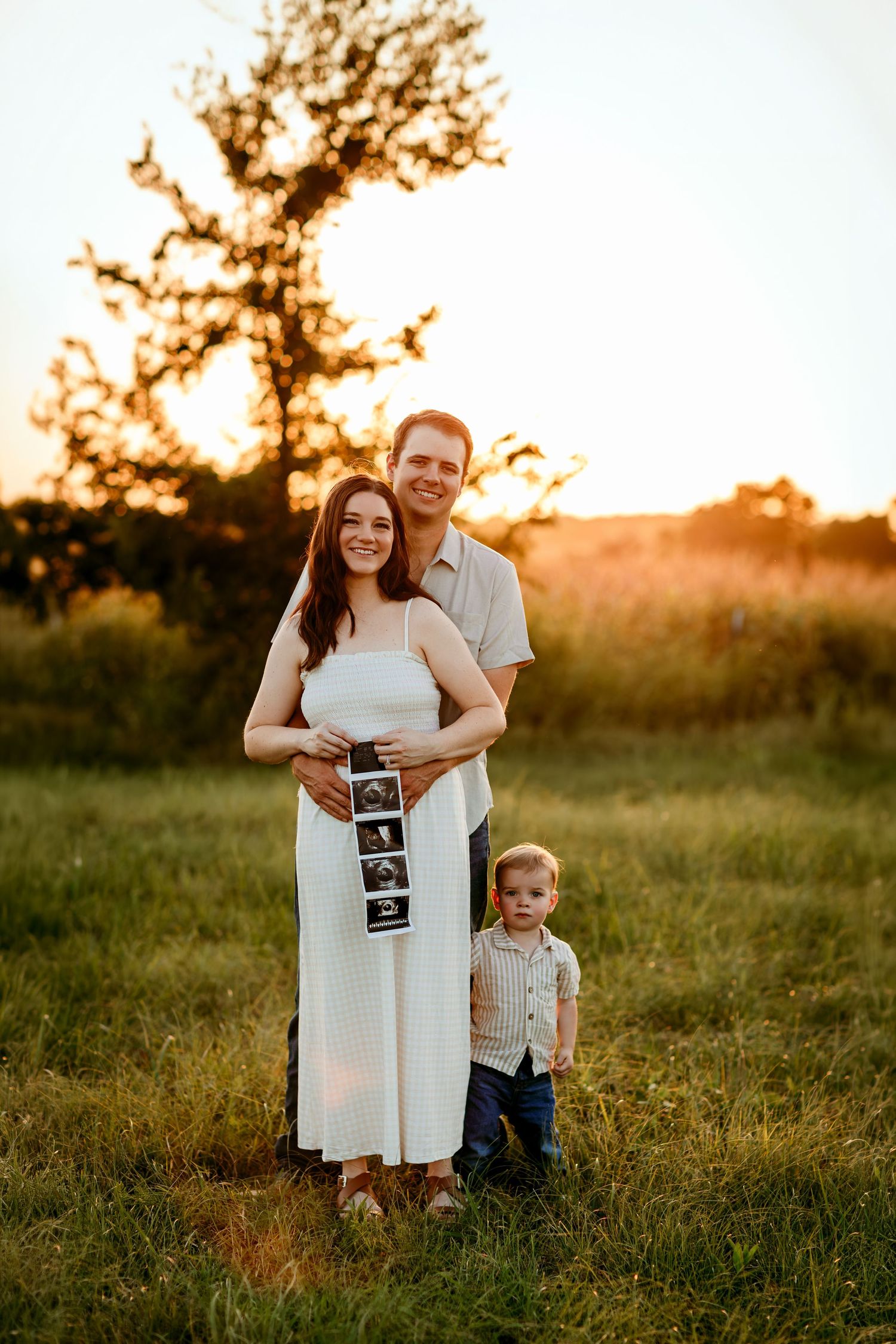 Family maternity photo at golden hour in a field, couple holding ultrasound images with toddler son.
