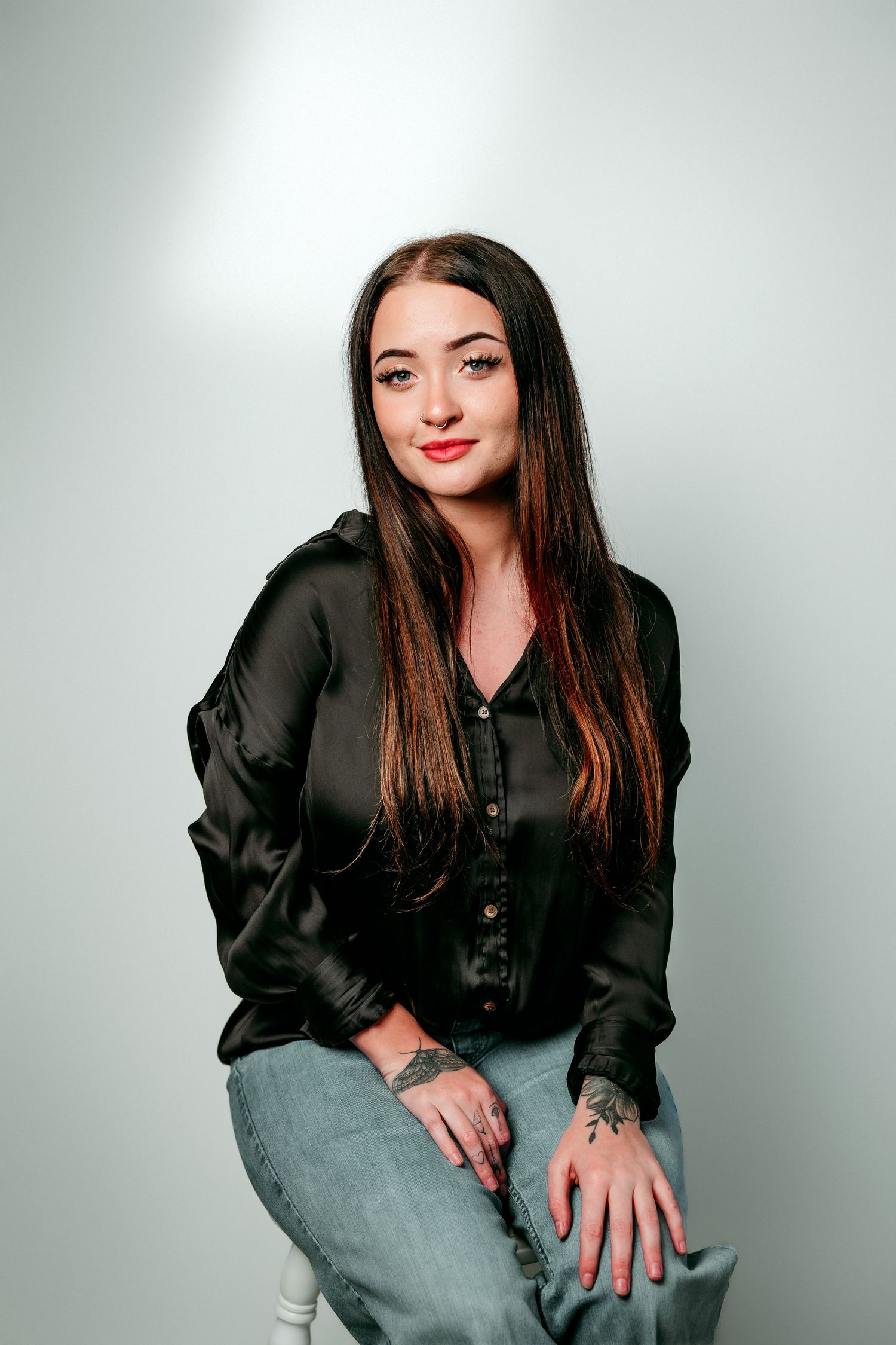 Young woman with long brown hair, red lips, black satin blouse, jeans, and hand tattoos sits against gray background.