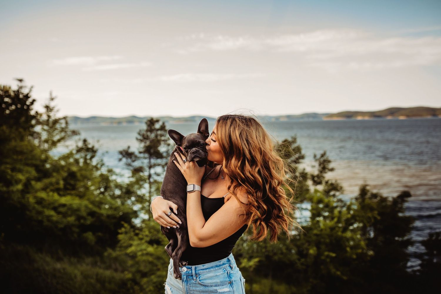 Young woman with long wavy hair kissing her French Bulldog outdoors near a scenic lake and green trees at sunset.