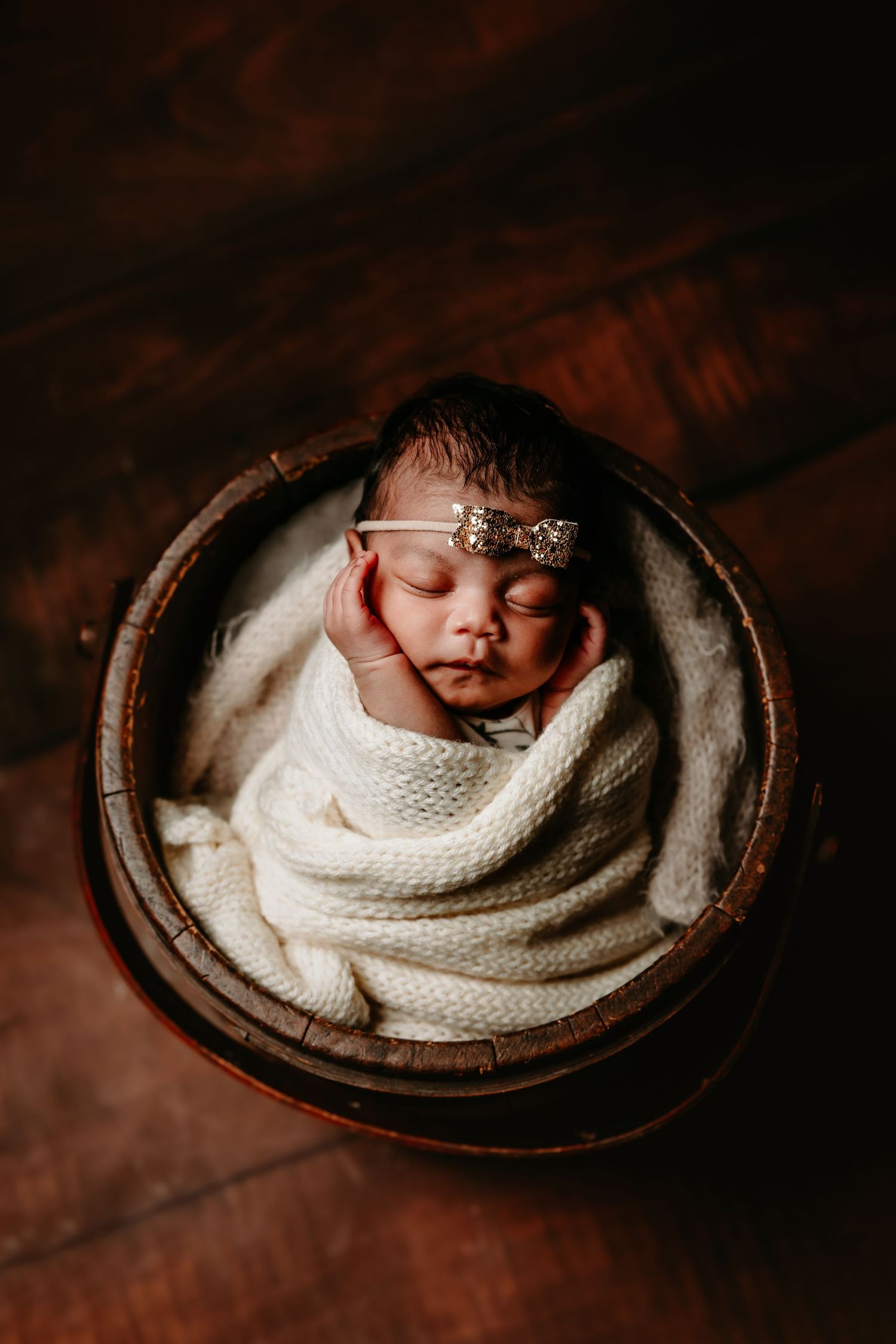 A newborn baby wrapped in white knit blanket sleeps peacefully in a rustic wooden bowl against dark background in a Paris, Texas studio.