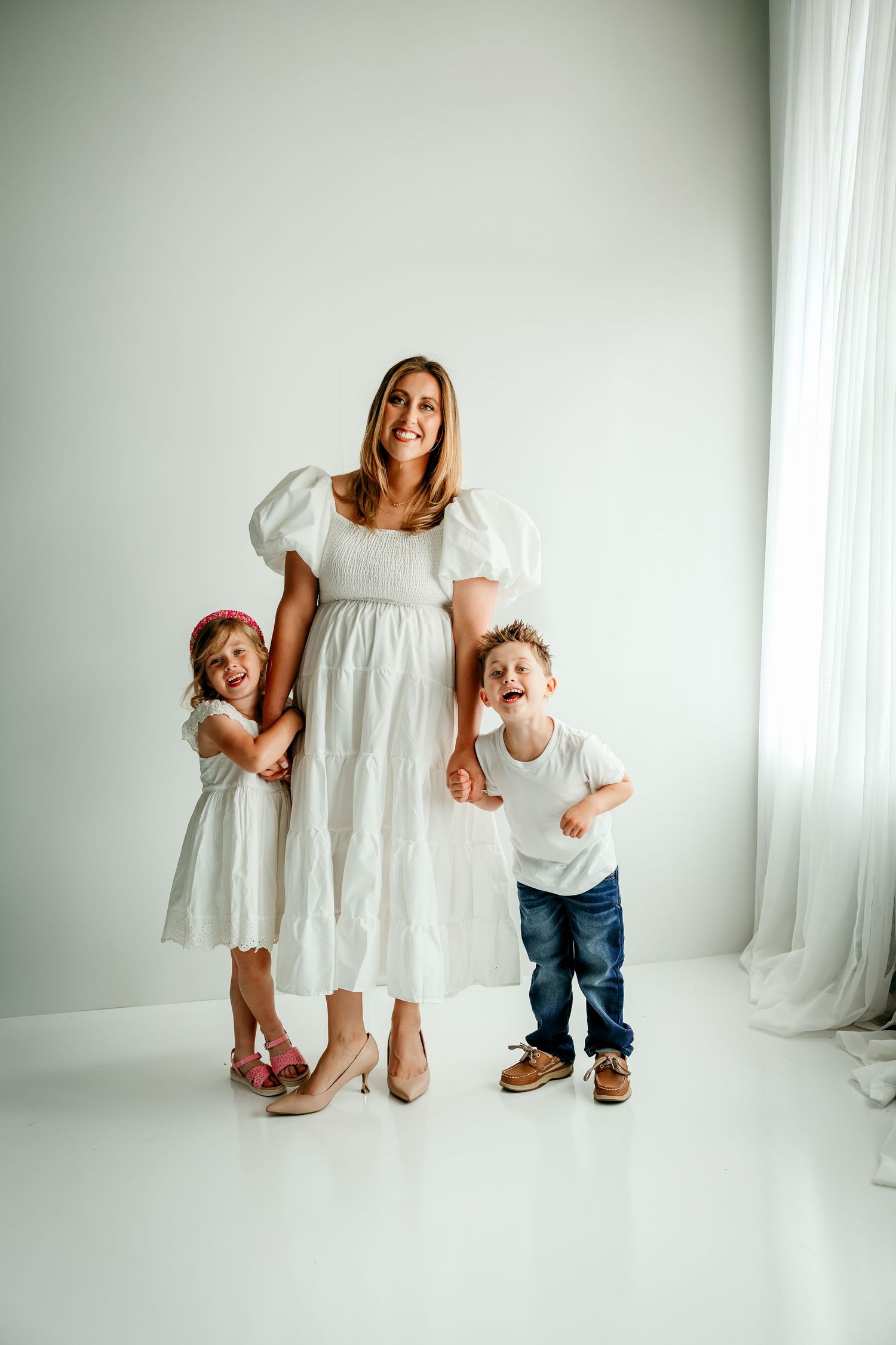 Pregnant woman in white dress posing with two young children in bright white studio setting.