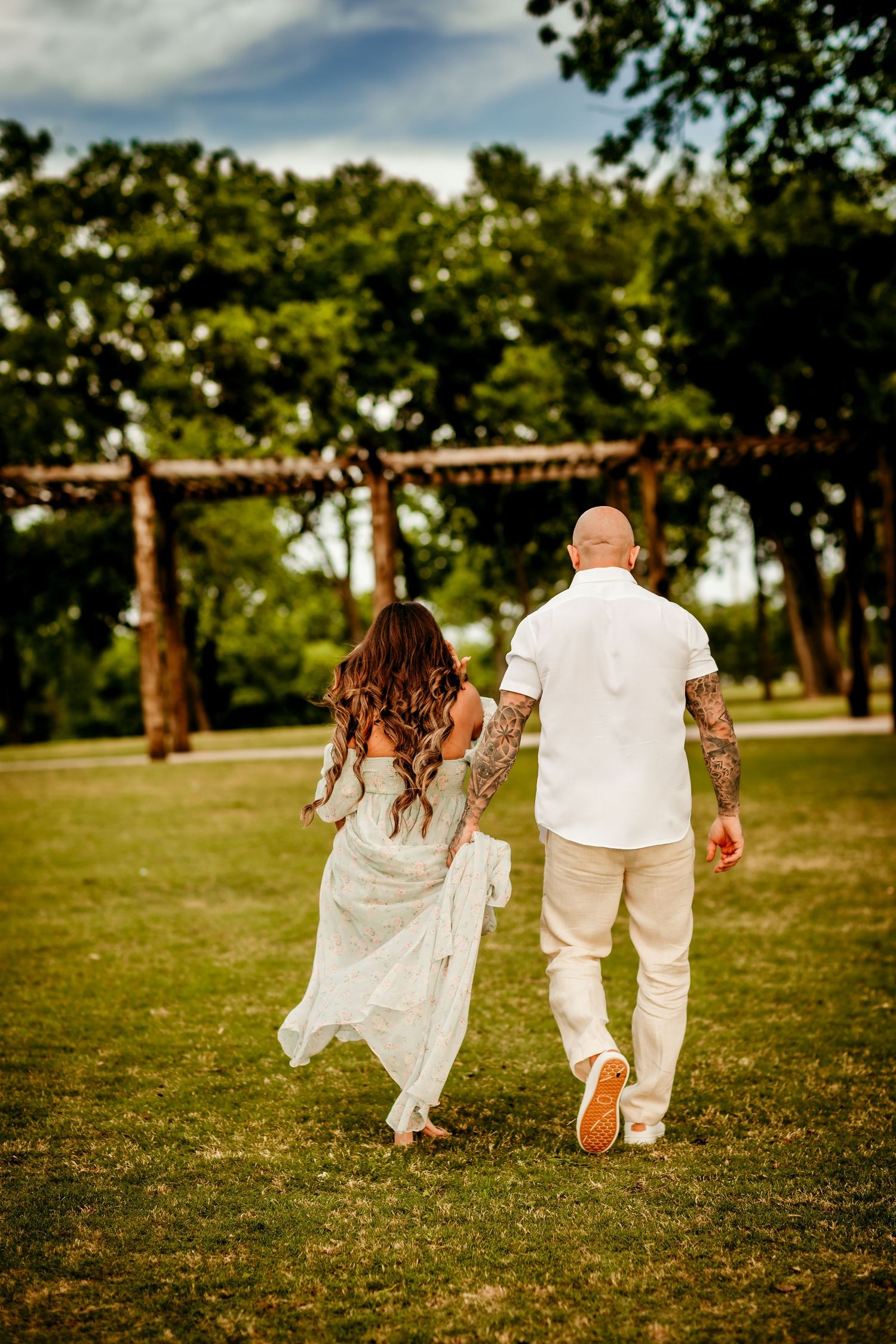 Tattooed couple in white attire walking hand-in-hand through a lush green park, viewed from behind.