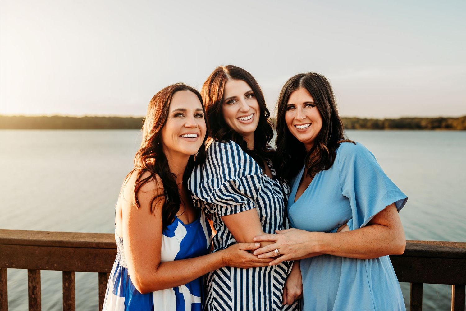 Three smiling women in blue dresses pose together on a wooden deck overlooking a lake at golden hour.