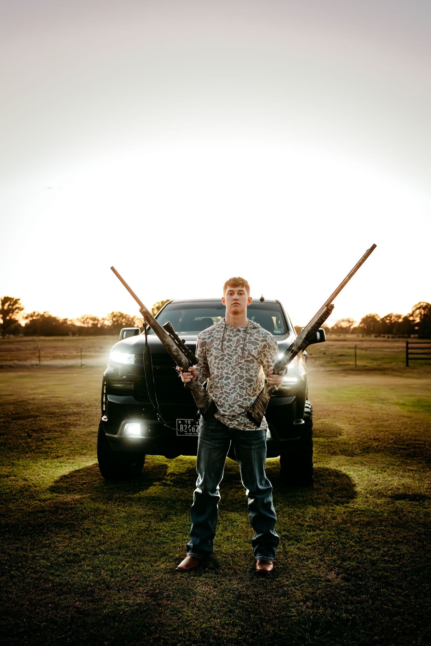 Young man holding two rifles in front of a truck at sunset on a rural farm property.
