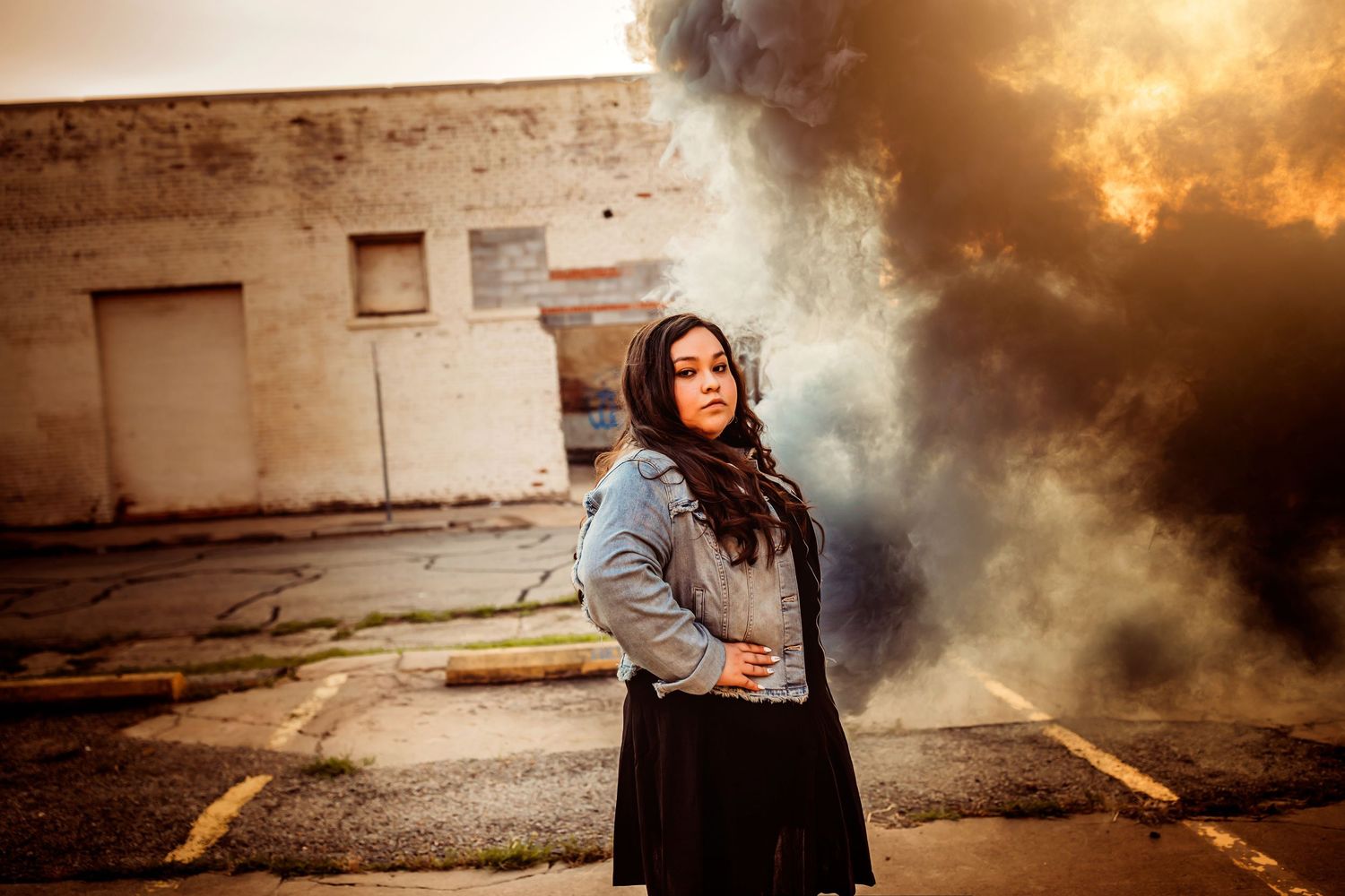 Woman in denim jacket and black skirt poses dramatically amid colorful smoke clouds outside an urban building.