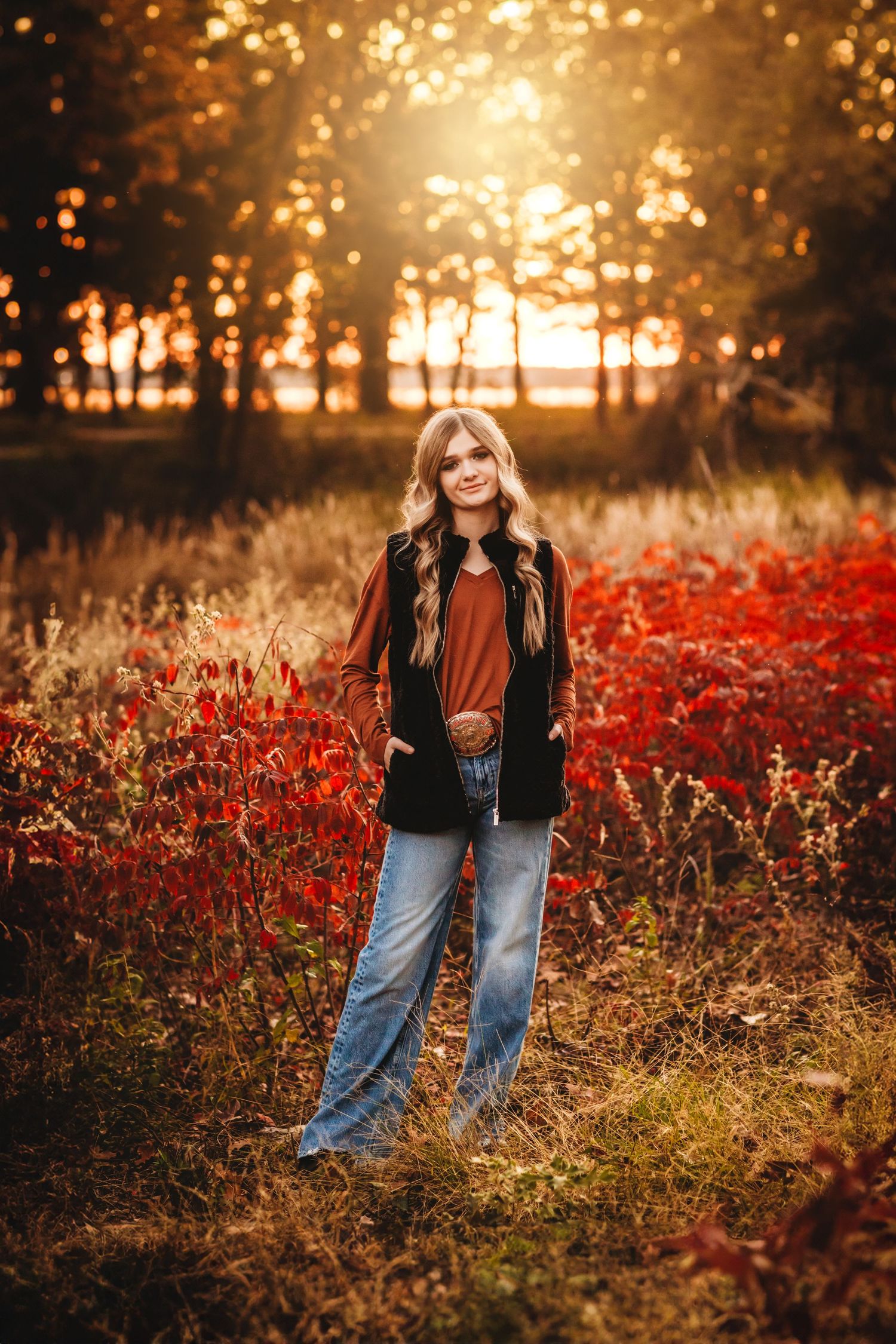 Young woman in rust top, black vest, and jeans poses in autumn field with red foliage and golden sunset bokeh.