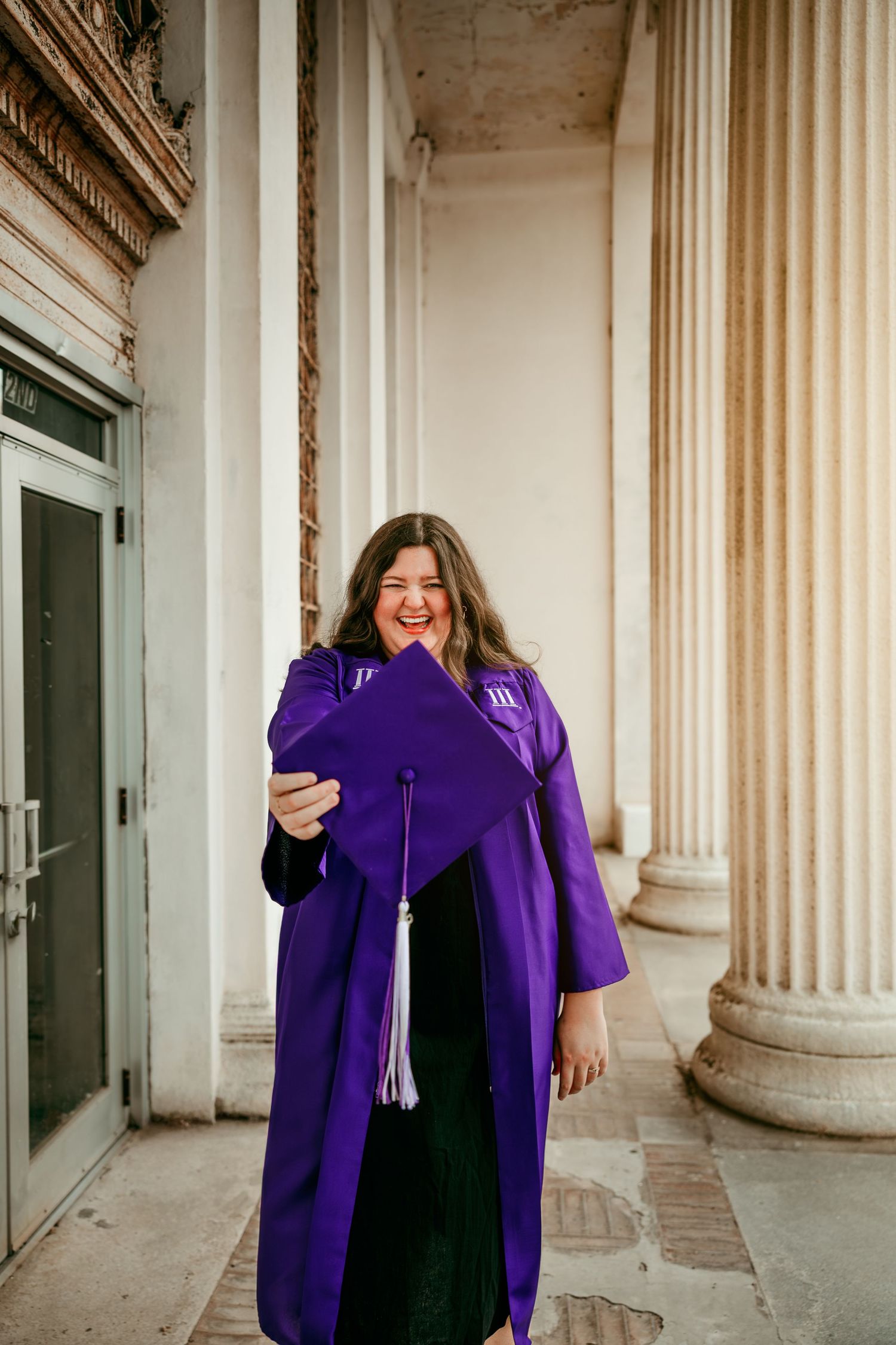 A joyful graduate in purple cap and gown holds her mortarboard outside a building with classical columns.