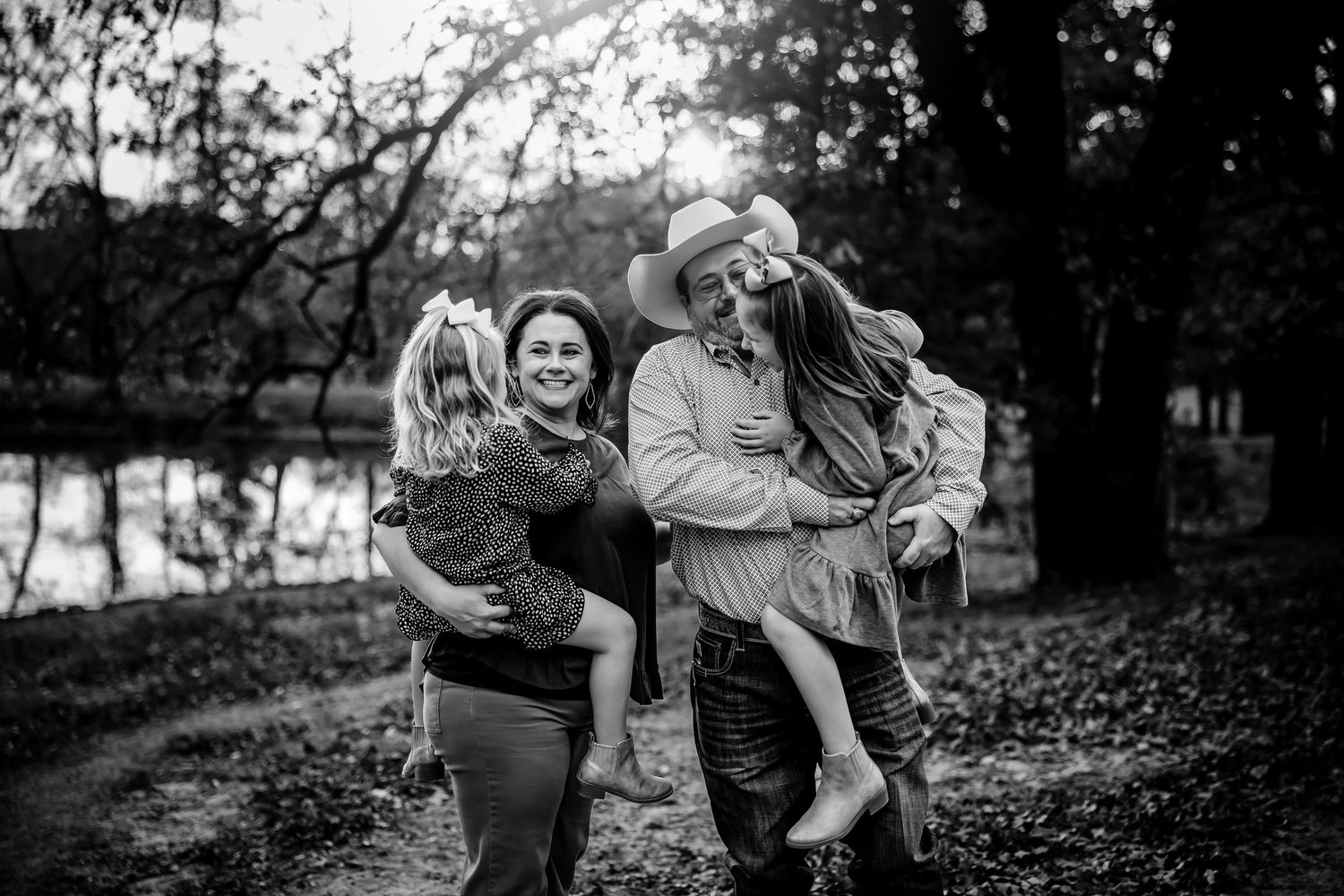 Black and white photo of a joyful family with two children outdoors near a pond surrounded by autumn trees.
