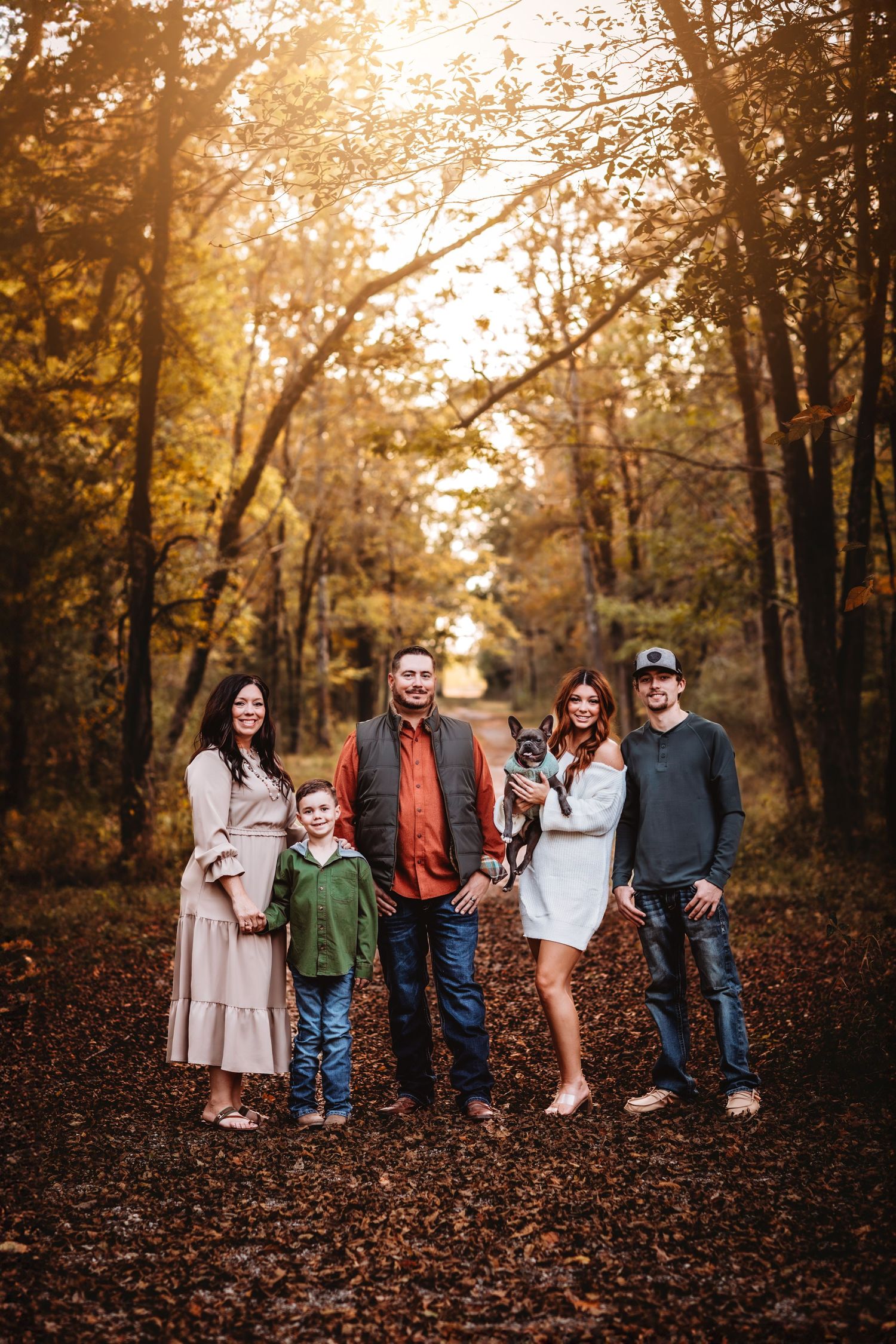 Family of five posing with a French Bulldog on a leaf-covered path surrounded by golden autumn trees.