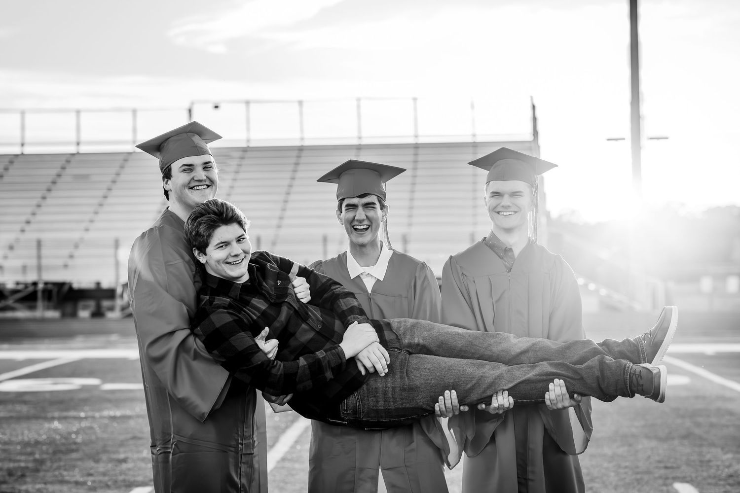 Four high school graduates in caps and gowns playfully carry a friend on a sunlit football field in black and white.