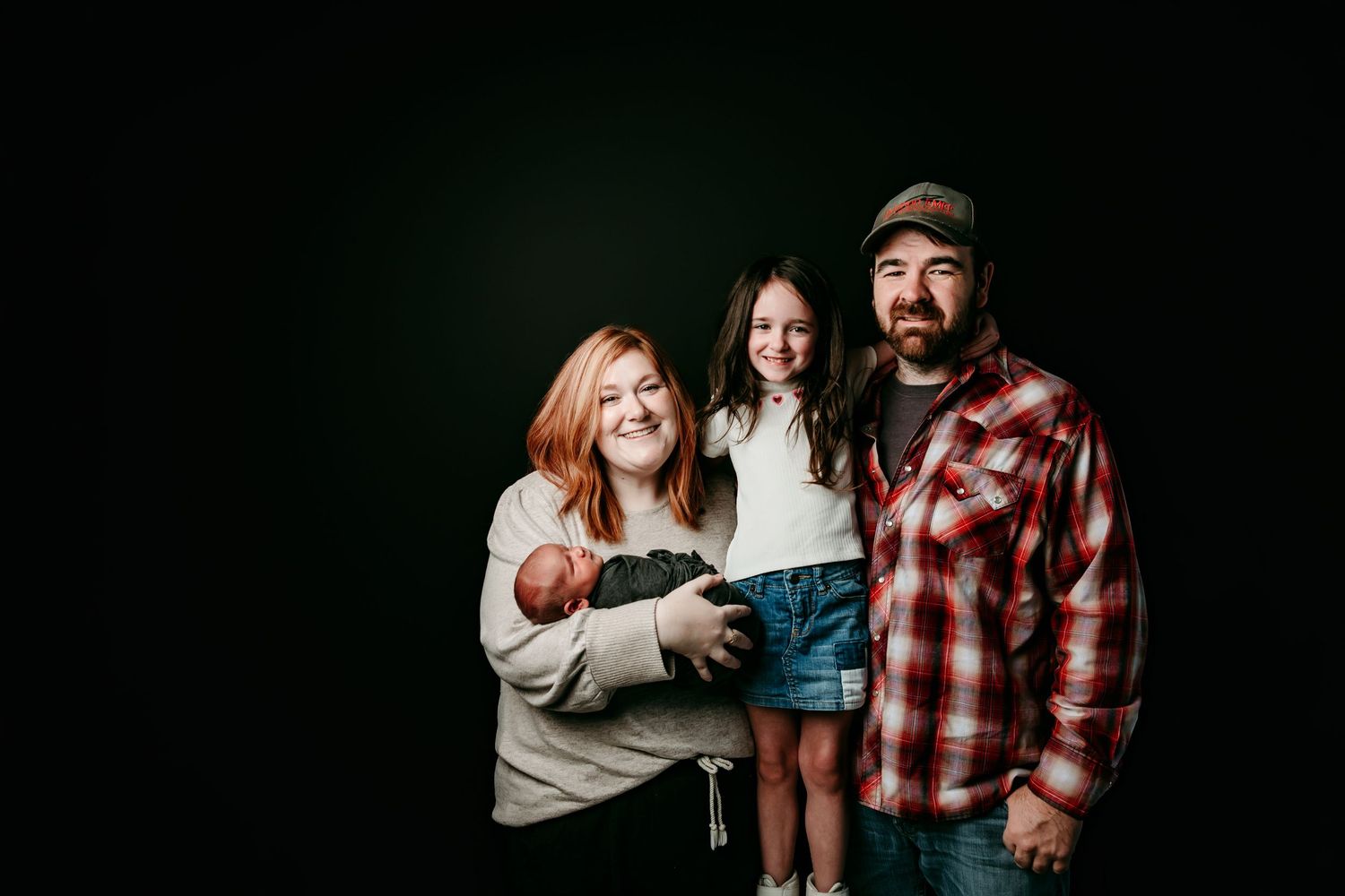 Family portrait of smiling parents with newborn baby and young girl against dark studio background.