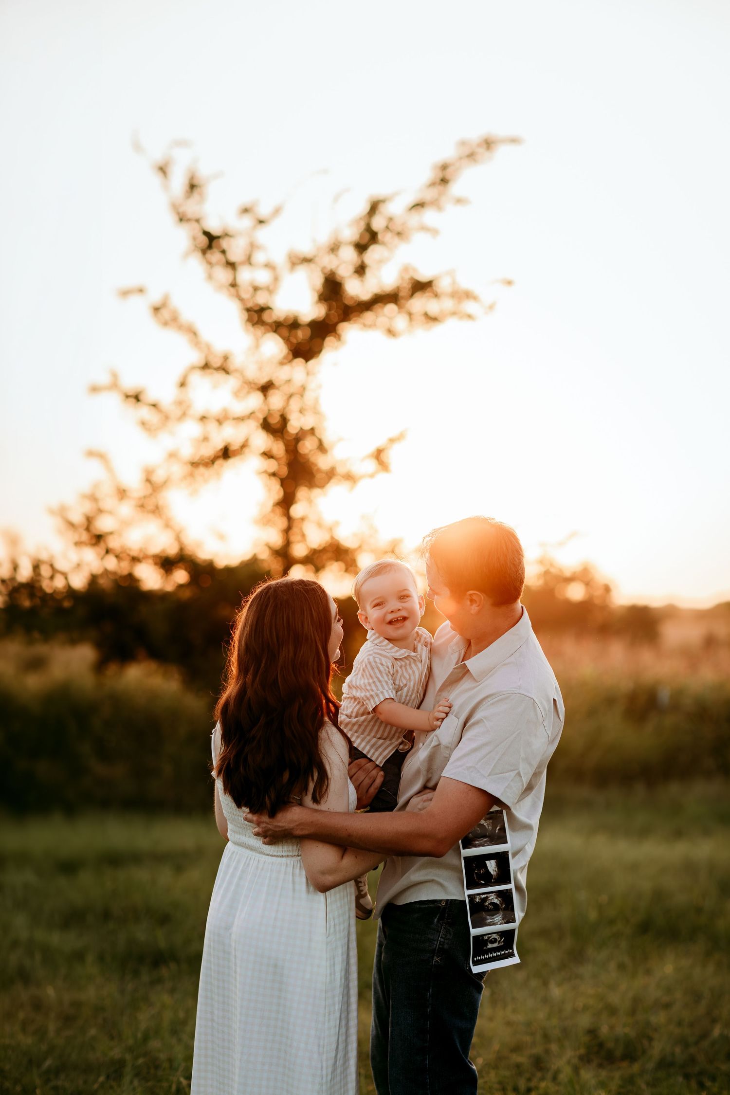 Family of three at golden hour, parents holding laughing baby with ultrasound photos, announcing second pregnancy outdoors.