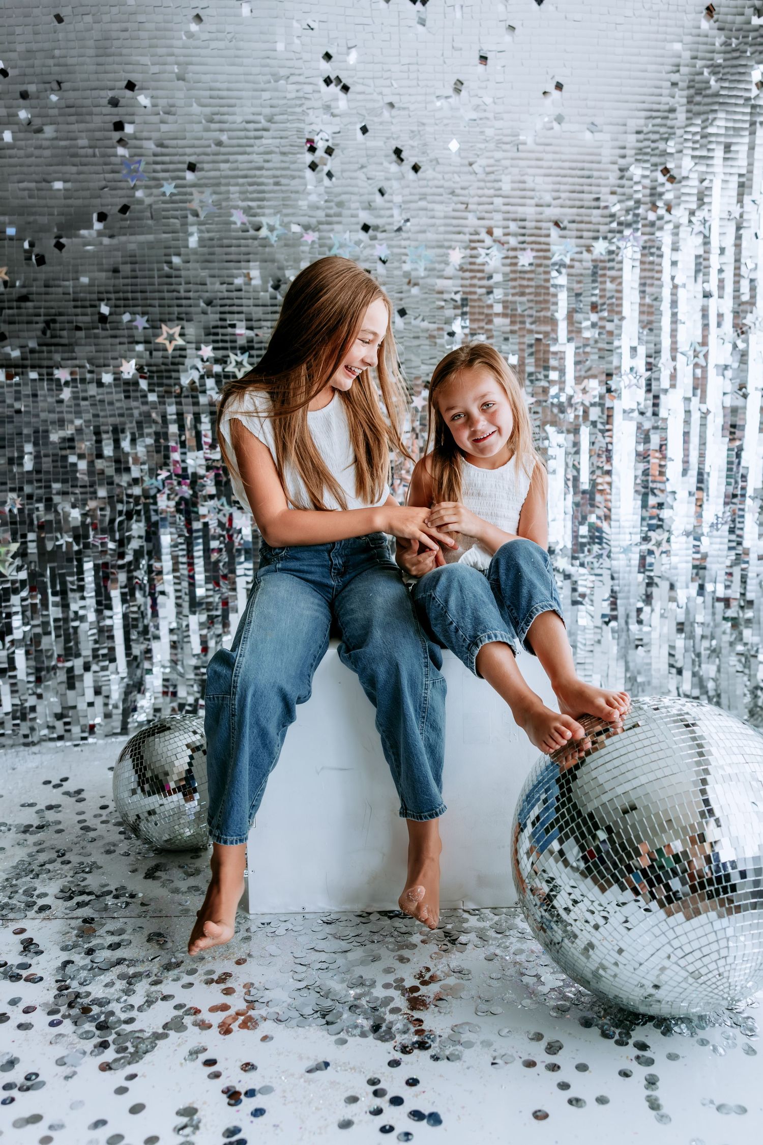 Two girls in jeans and white tops laugh together surrounded by disco balls and silver confetti at a party.
