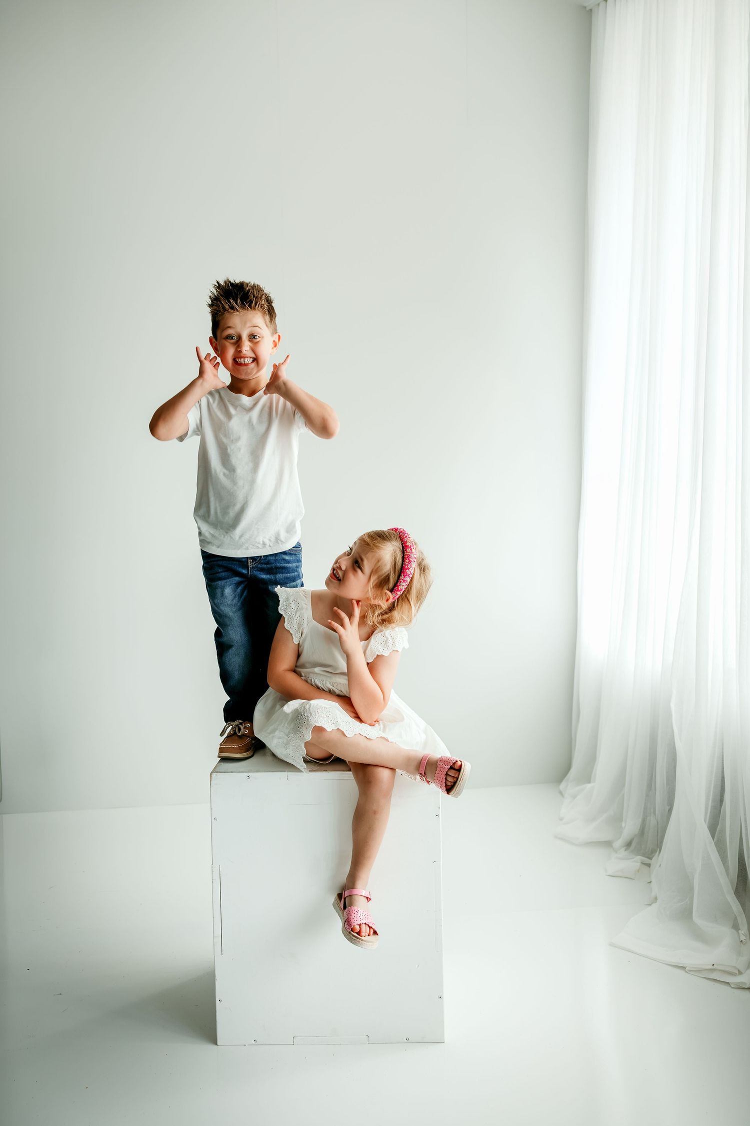Two playful children posing in a white studio, boy making funny faces standing on a cube, girl sitting elegantly in a dress.