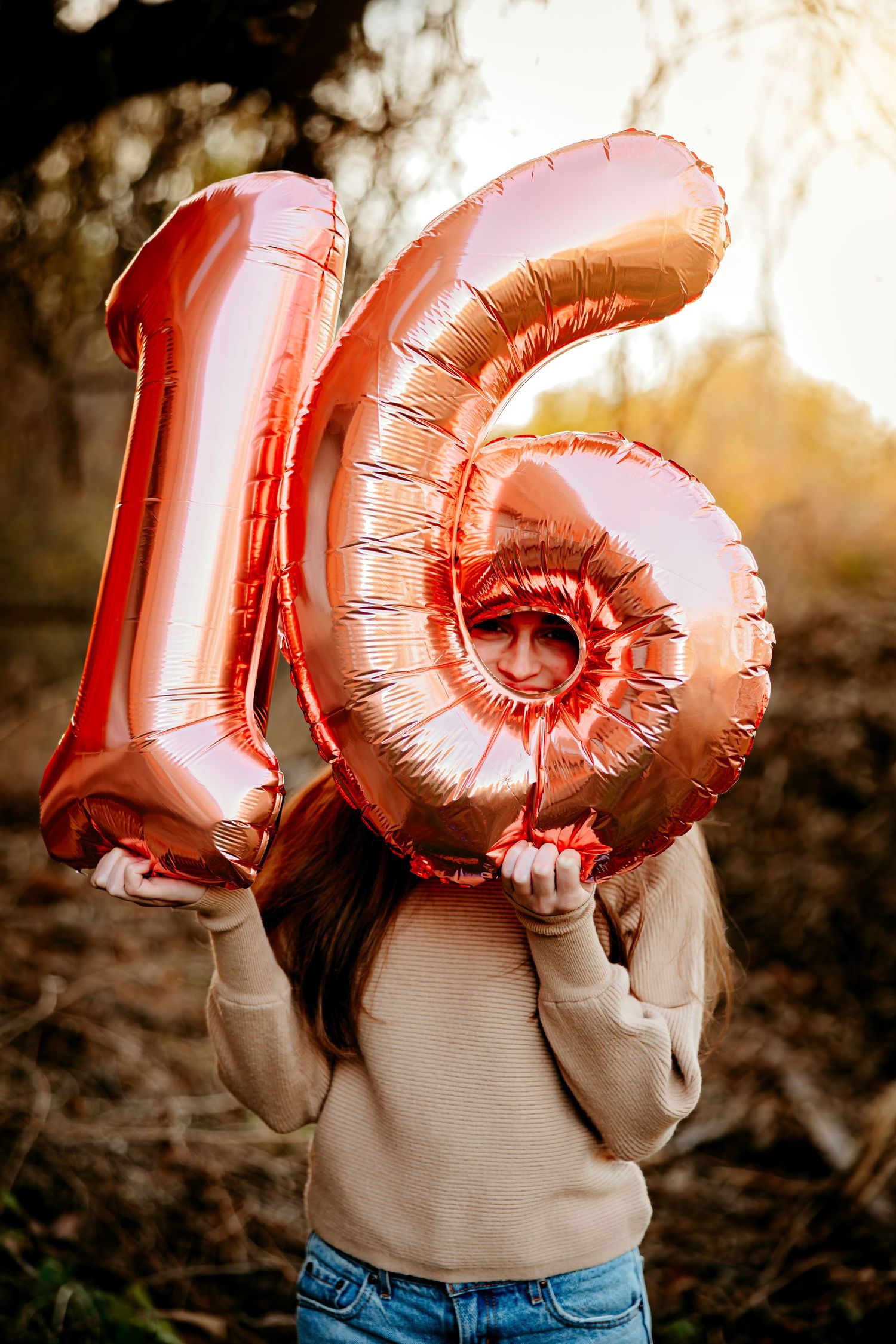 Person holding rose gold number 16 foil balloons outdoors at sunset in casual attire for a sweet 16 photo session in Paris, Texas.