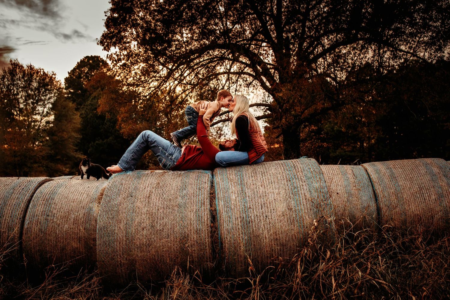 Couple sitting on hay bales at sunset in a rustic field surrounded by autumn trees in warm golden light.