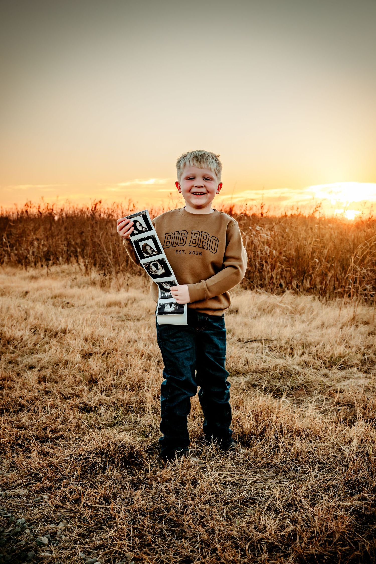 A smiling boy holds ultrasound photos in a field at sunset, wearing a Big Bro sweatshirt announcing a new sibling.