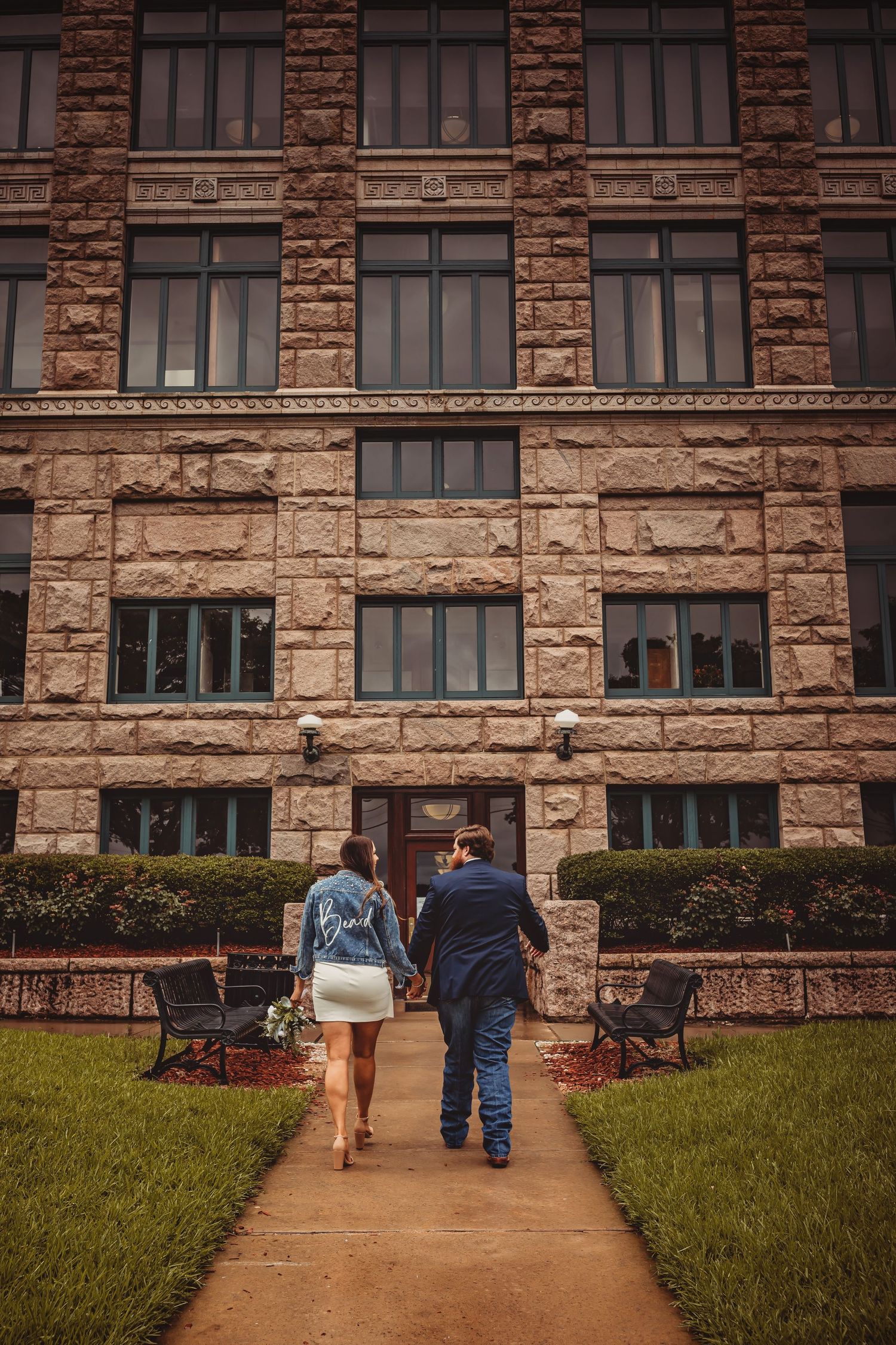 Couple walking hand-in-hand toward a historic stone building entrance, woman wearing a denim jacket with jeans and white dress.