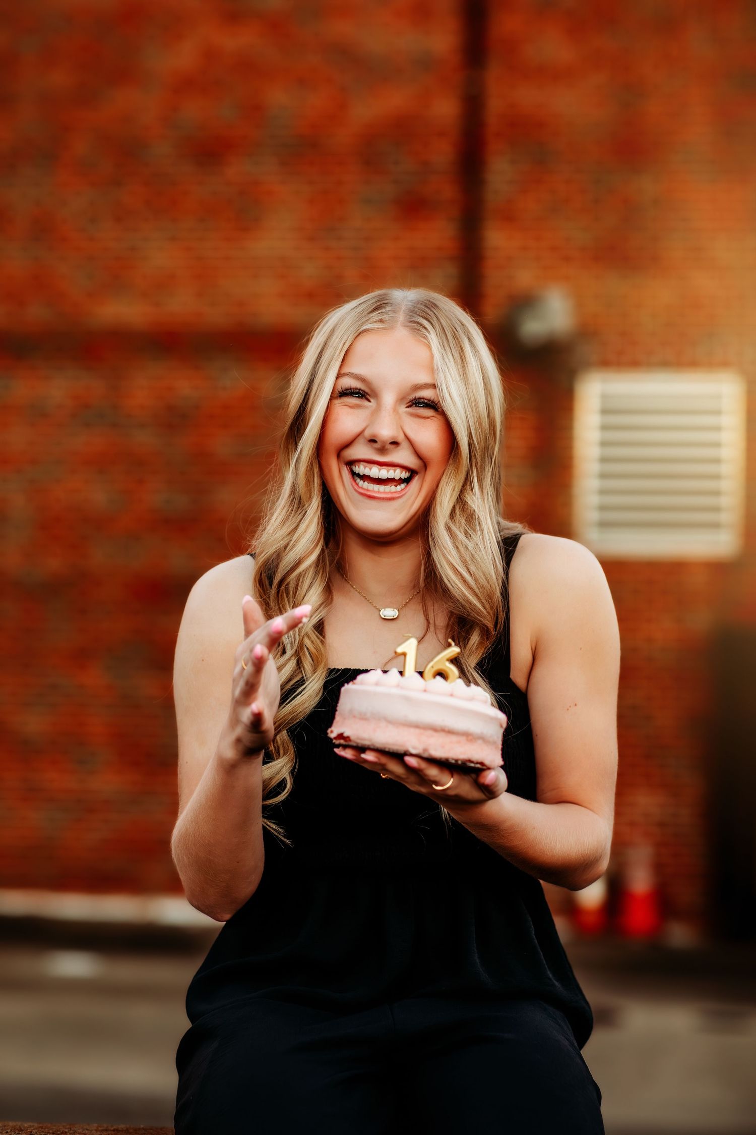 Smiling blonde woman holding a pink birthday cake with gold number 16 candles, wearing a black dress outdoors.