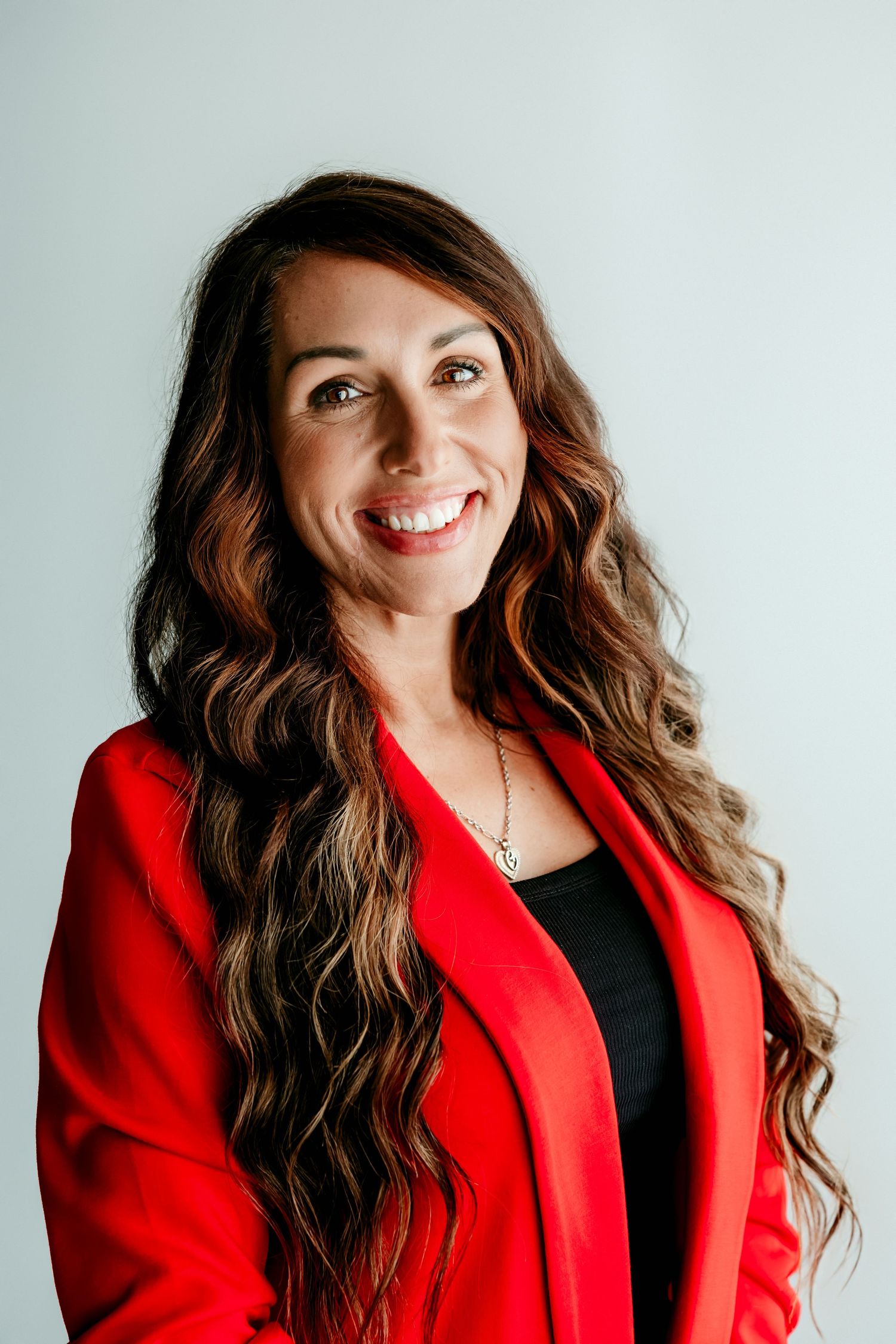 Professional woman in red blazer with long wavy brown hair smiling confidently against light gray background.