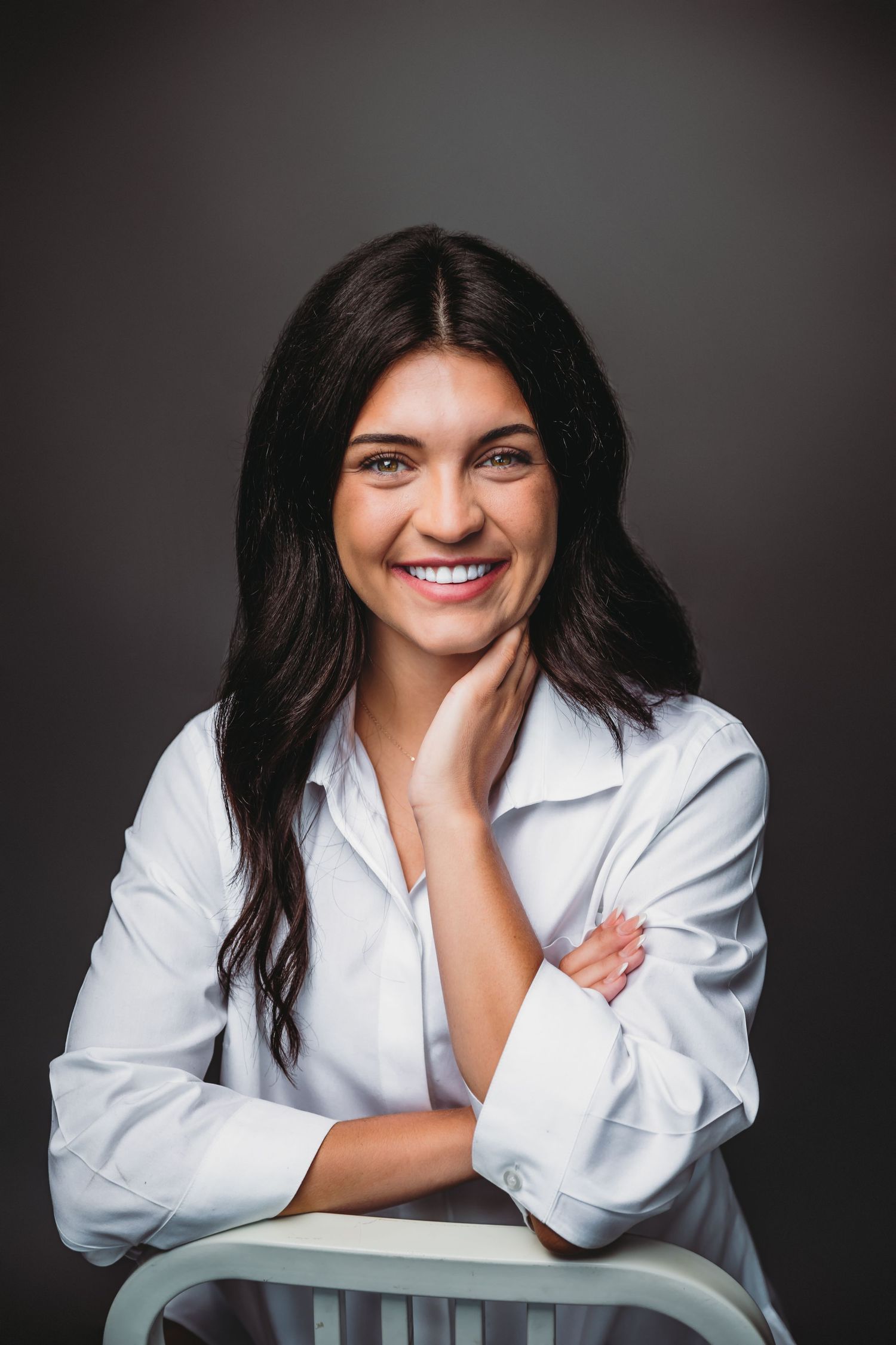 Smiling young woman with dark hair in white button-up shirt, posing confidently against dark gray background.