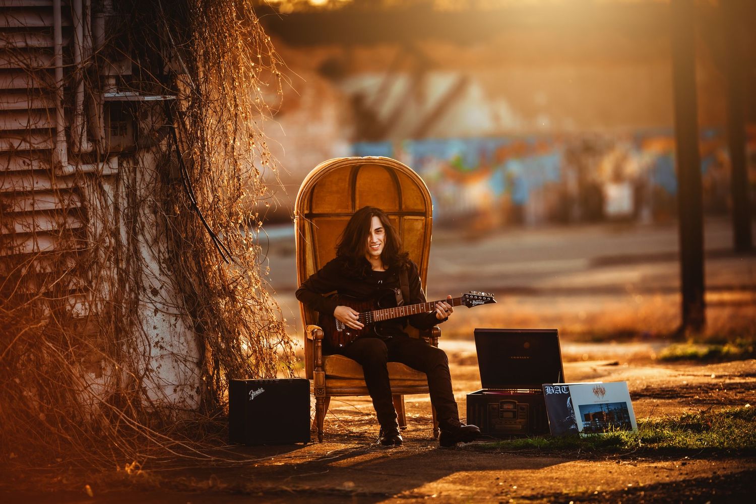 Musician playing electric guitar outdoors at golden hour, seated in a wicker chair near a tree on a sunlit street.
