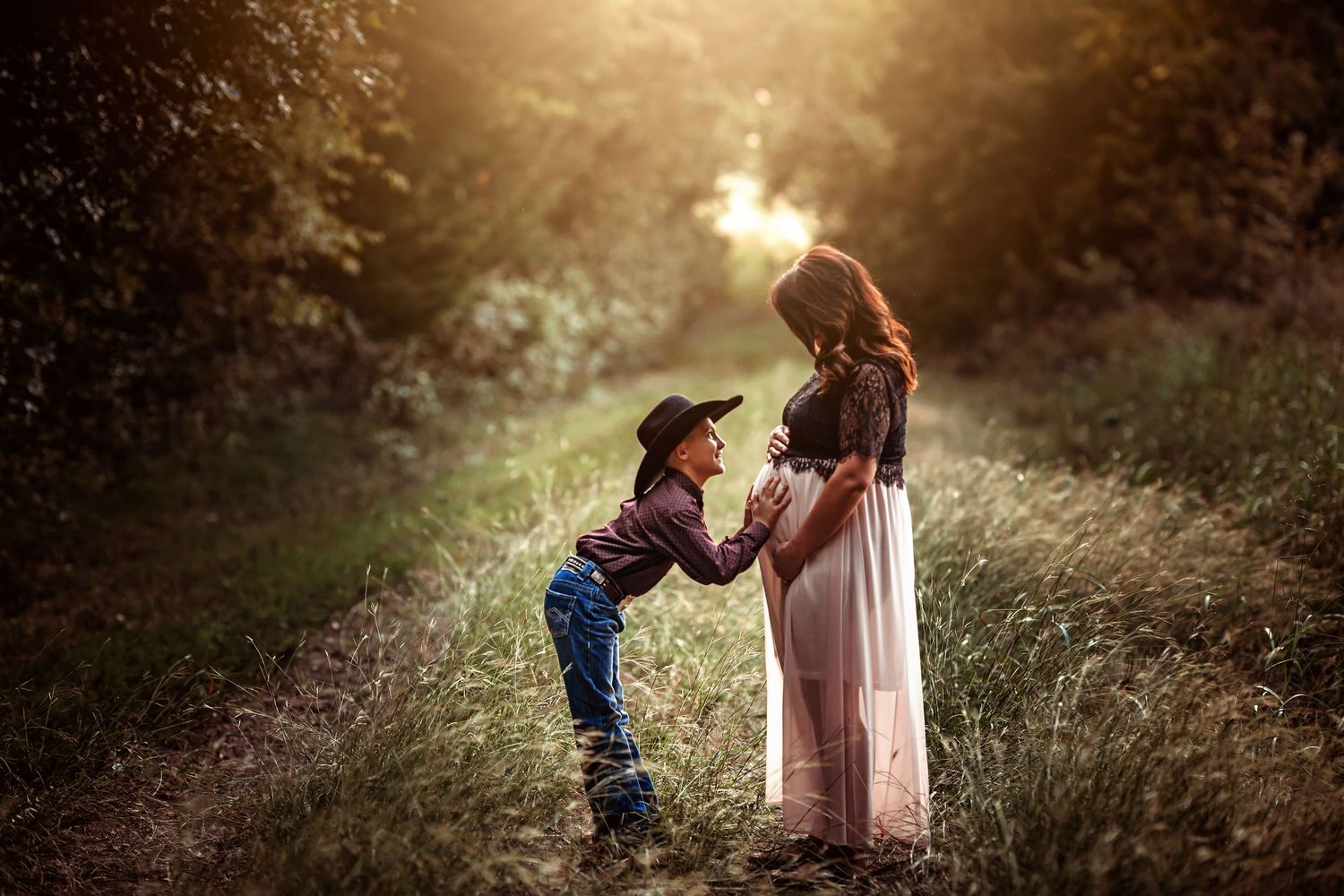 A young boy in a cowboy hat kneels before a pregnant woman in a sunlit forest path, holding her hands tenderly.