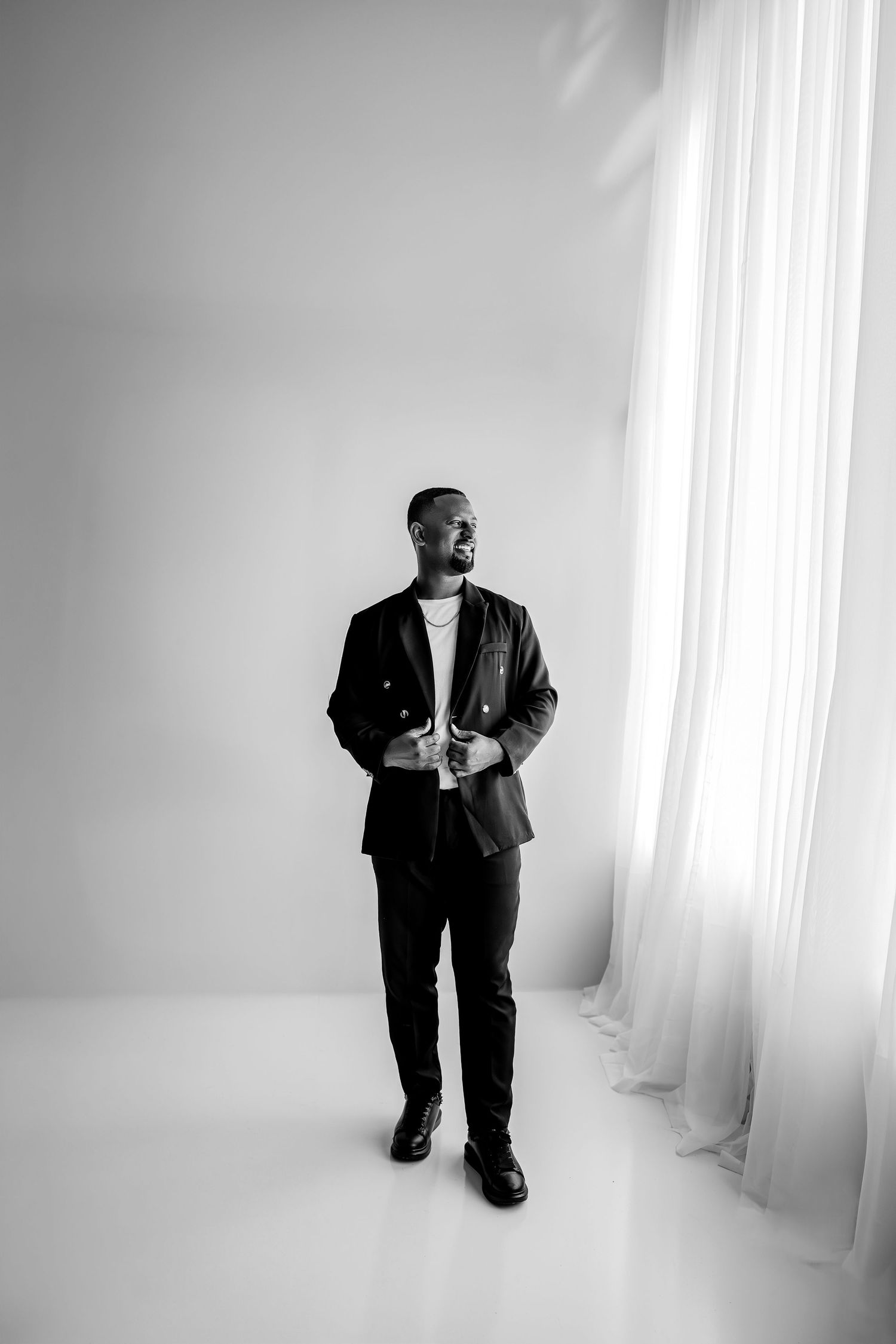 Black and white portrait of a stylish man in a dark blazer standing confidently near sheer white curtains.