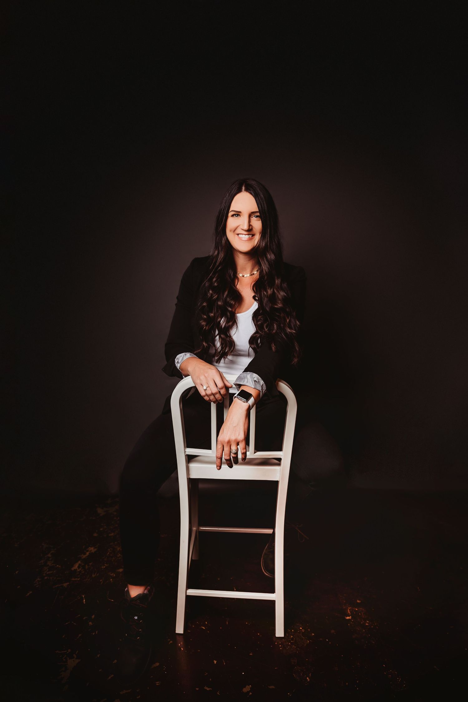 Woman in black blazer smiling while seated on white chair against dark studio background.