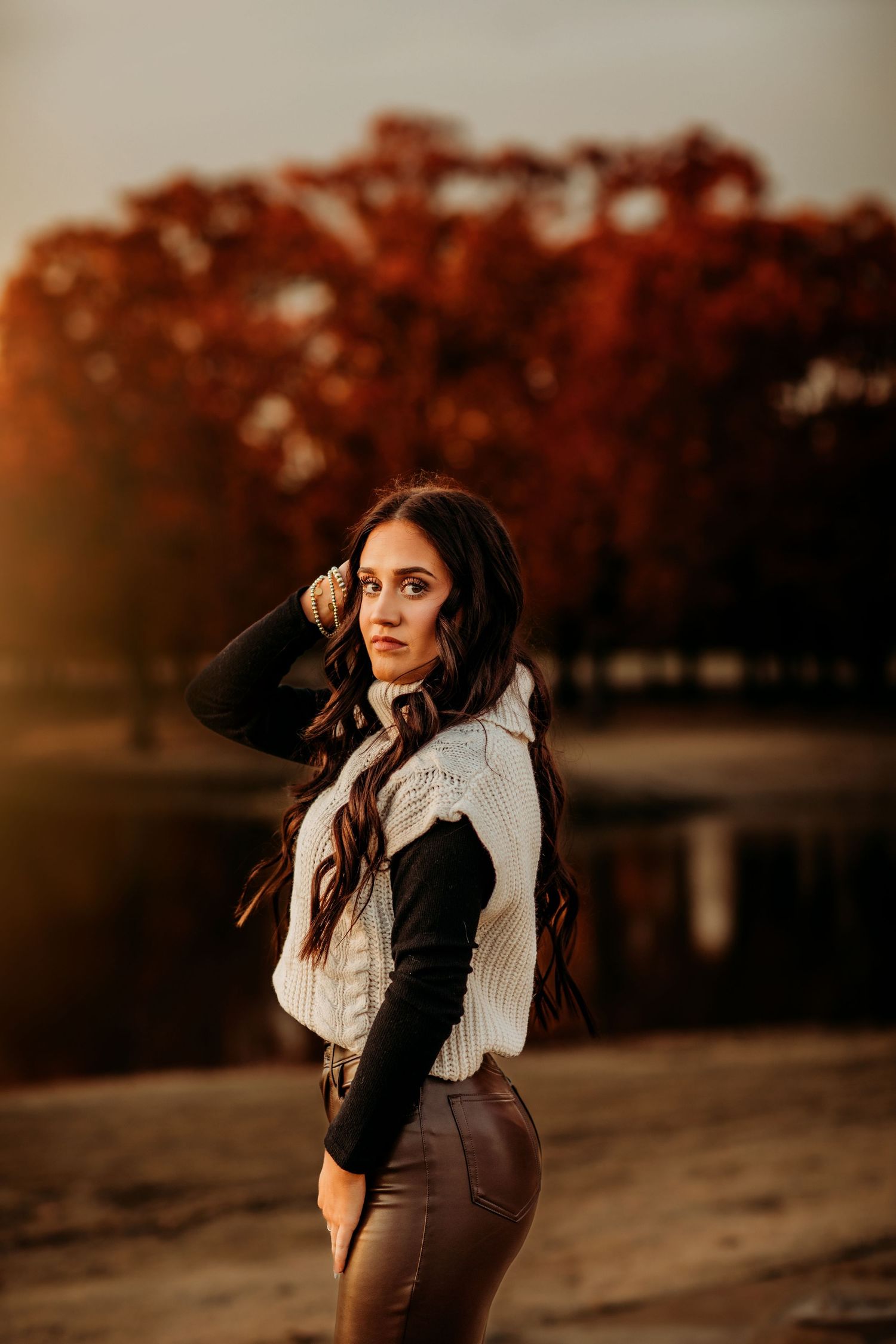 Young woman with long dark hair poses outdoors in autumn, wearing a white sweater and brown pants near a pond.