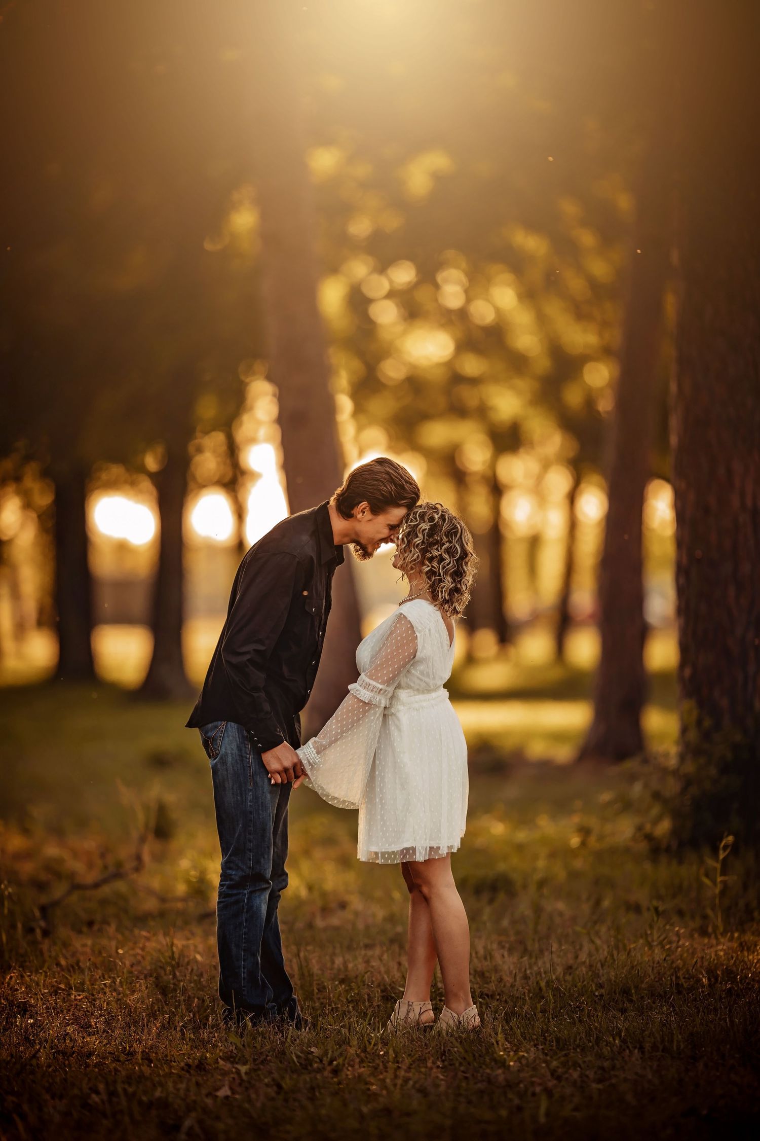 Romantic couple sharing a tender moment in a sunlit forest during golden hour, woman in white dress.