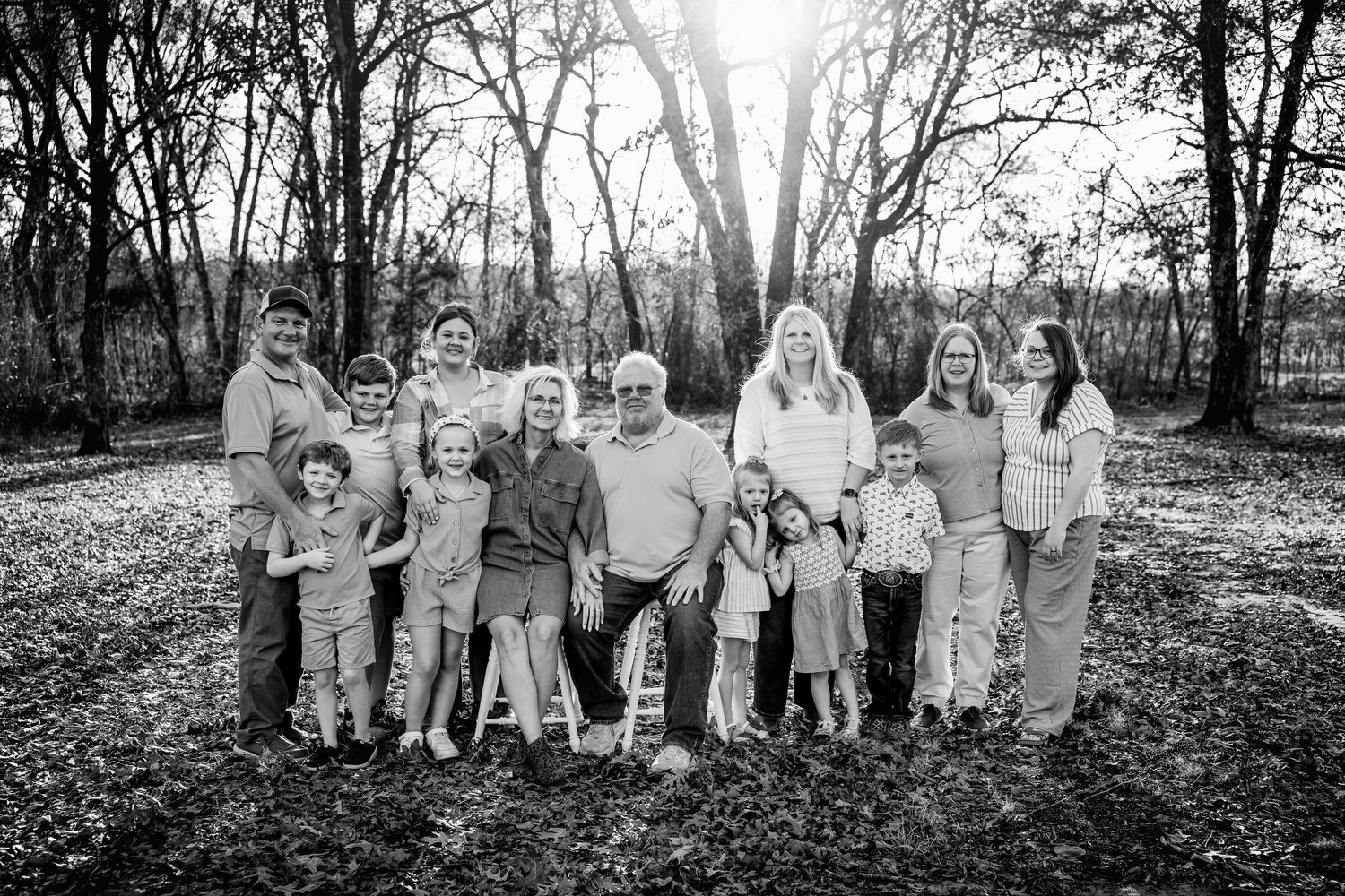 Black and white family portrait of multigenerational group posing outdoors among trees in natural woodland setting.