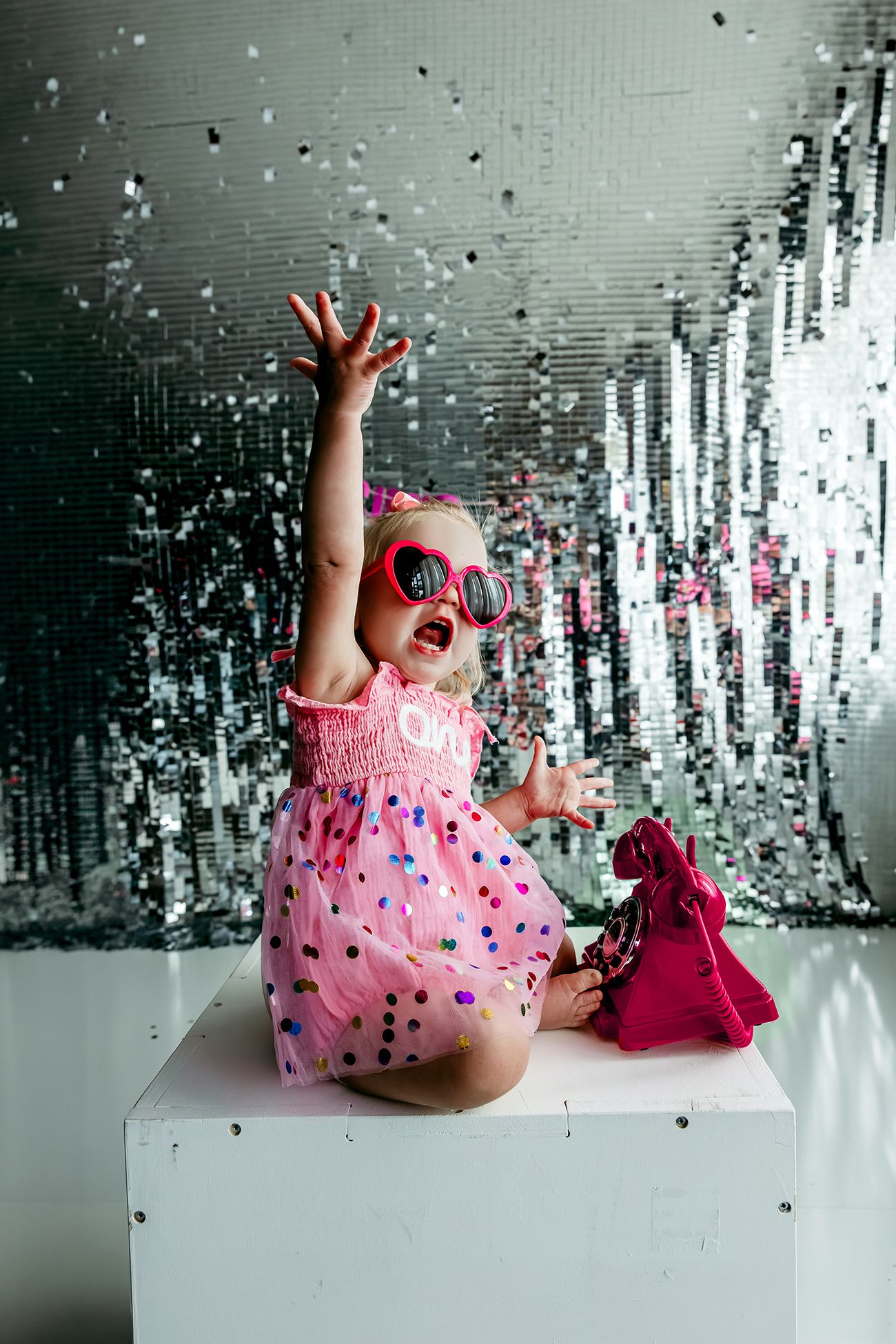 Joyful baby girl in pink polka dot dress and heart sunglasses reaching for confetti against silver sequin backdrop.