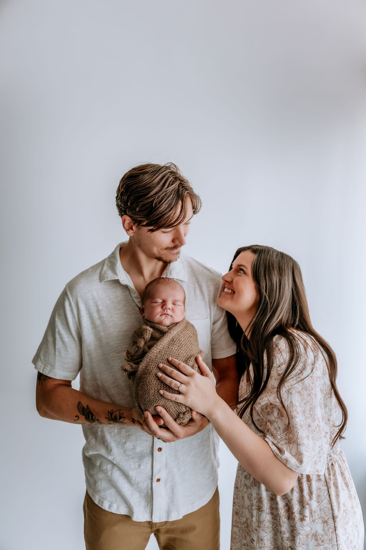 Young couple smiling at each other while holding their newborn baby wrapped in a brown swaddle blanket.