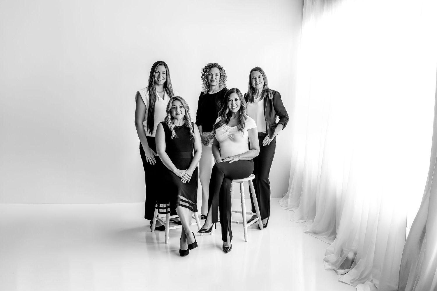 Black and white studio portrait of four professional women posing together, two standing and two seated, in elegant attire.