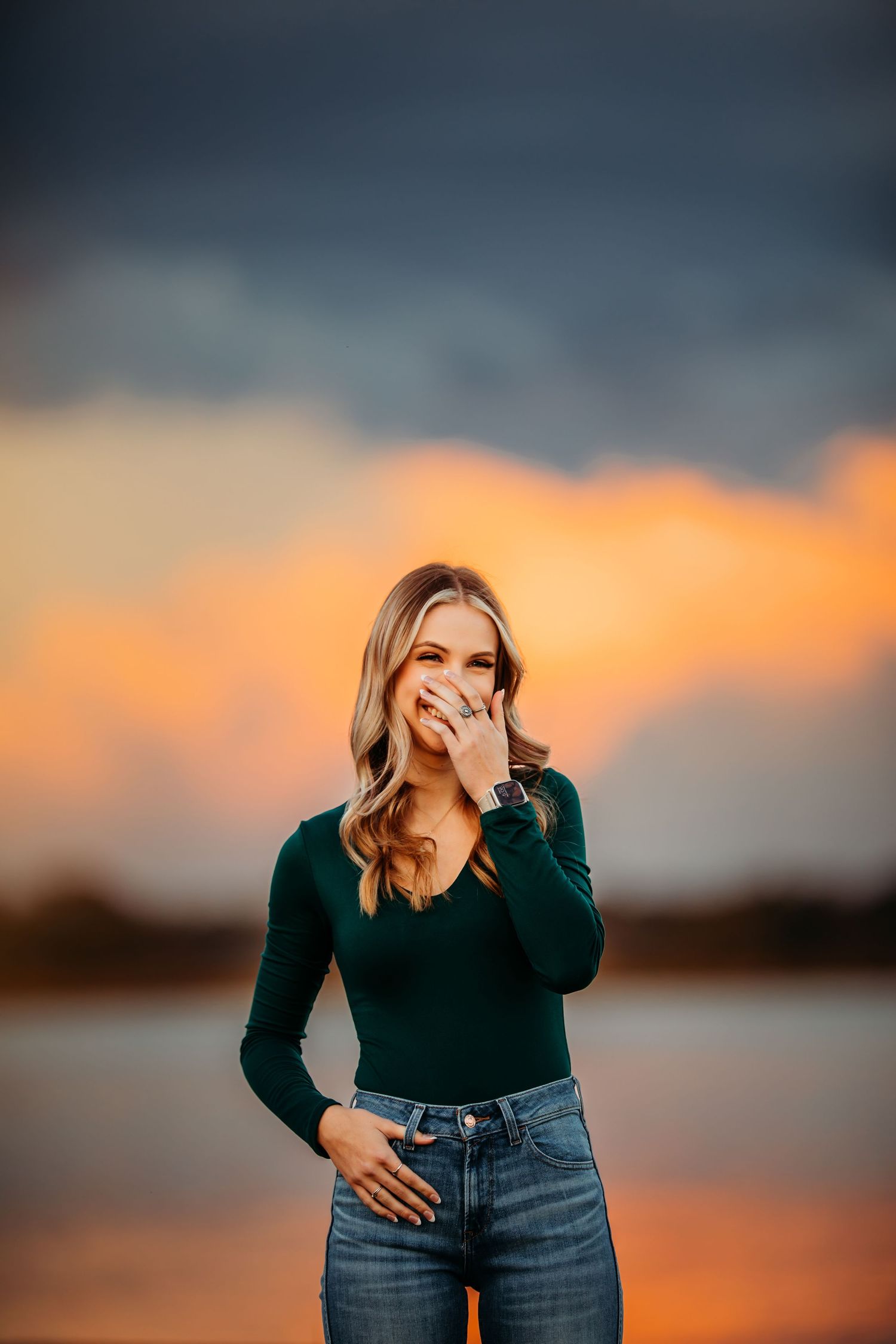 Young woman in green top and jeans laughing at sunset with dramatic orange clouds behind her.