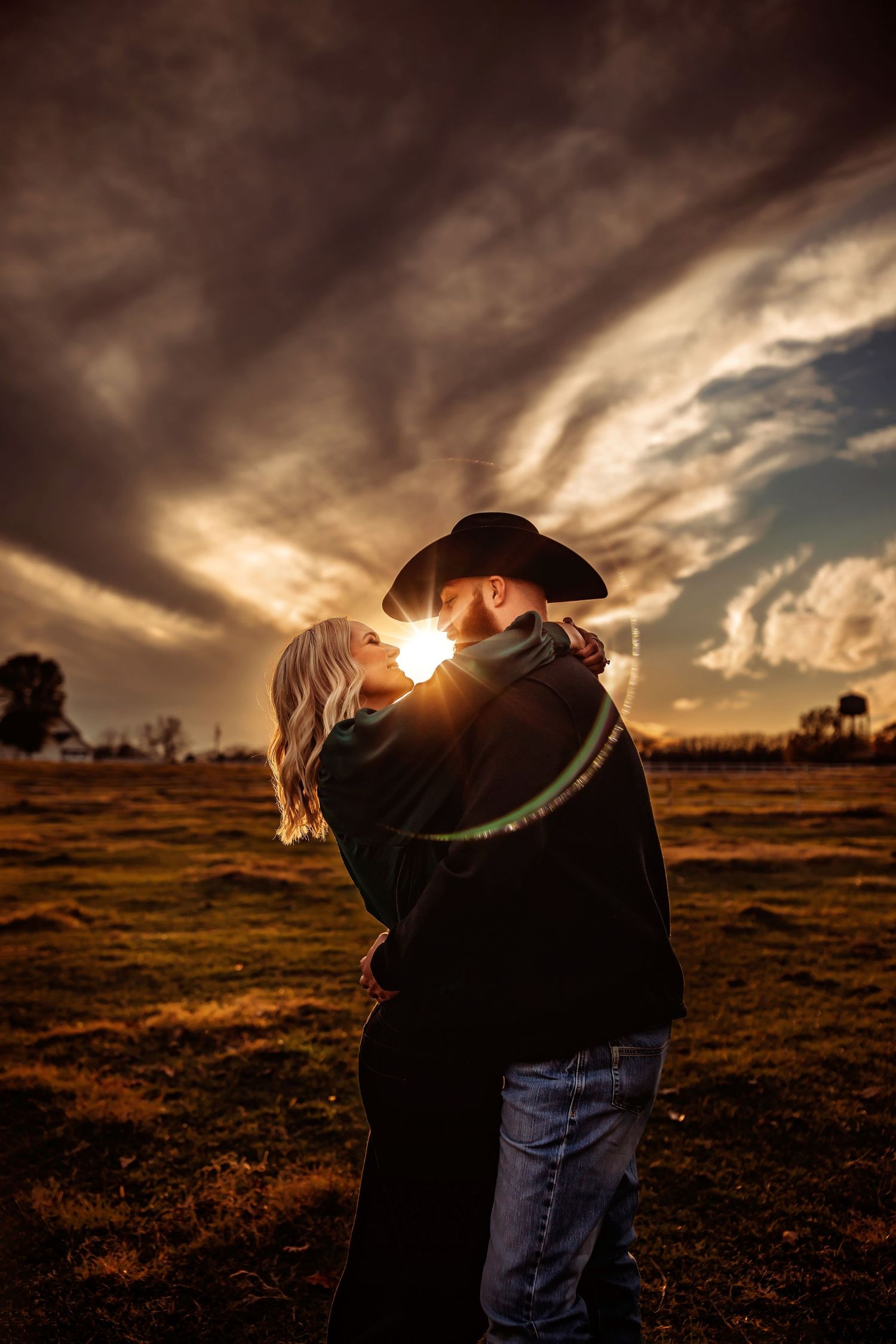 Couple embracing at golden sunset in a field, man in cowboy hat and woman with blonde hair, dramatic stormy sky backdrop.