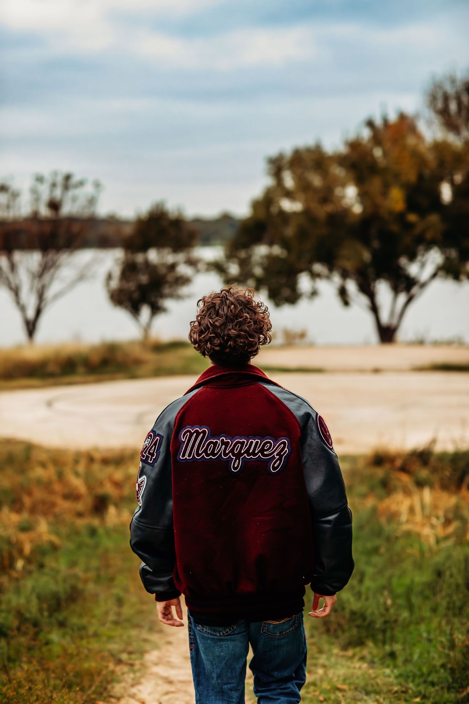 Person wearing a burgundy Marquez varsity jacket with leather sleeves standing on a rural path surrounded by trees.