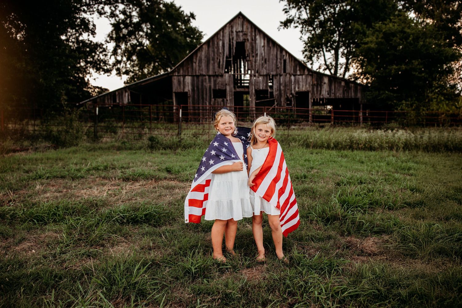 Two young girls wrapped in American flags stand in a green field with a rustic wooden barn in the background.
