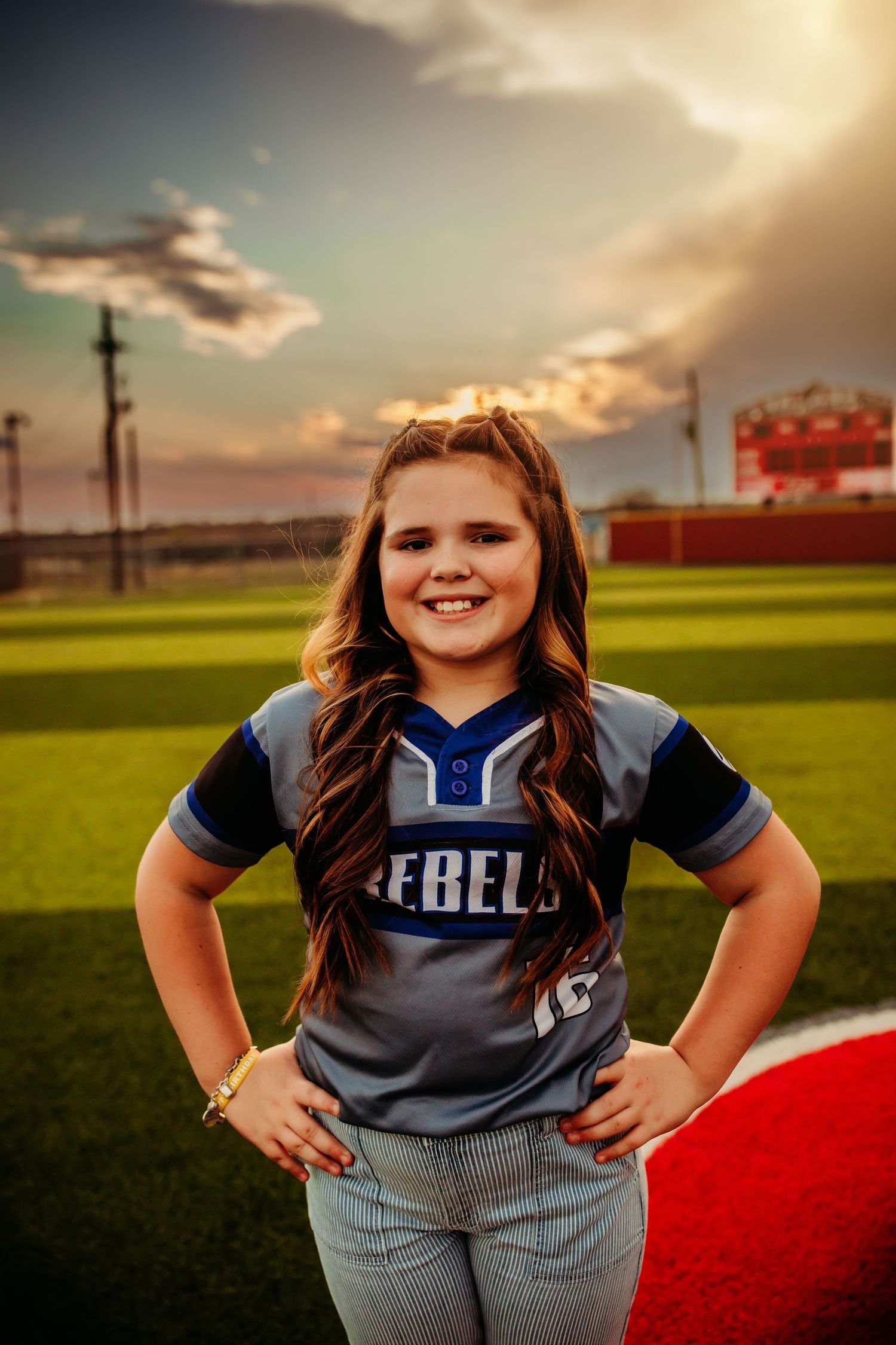 Young girl in Rebels softball jersey smiling on green field at sunset with hands on hips.