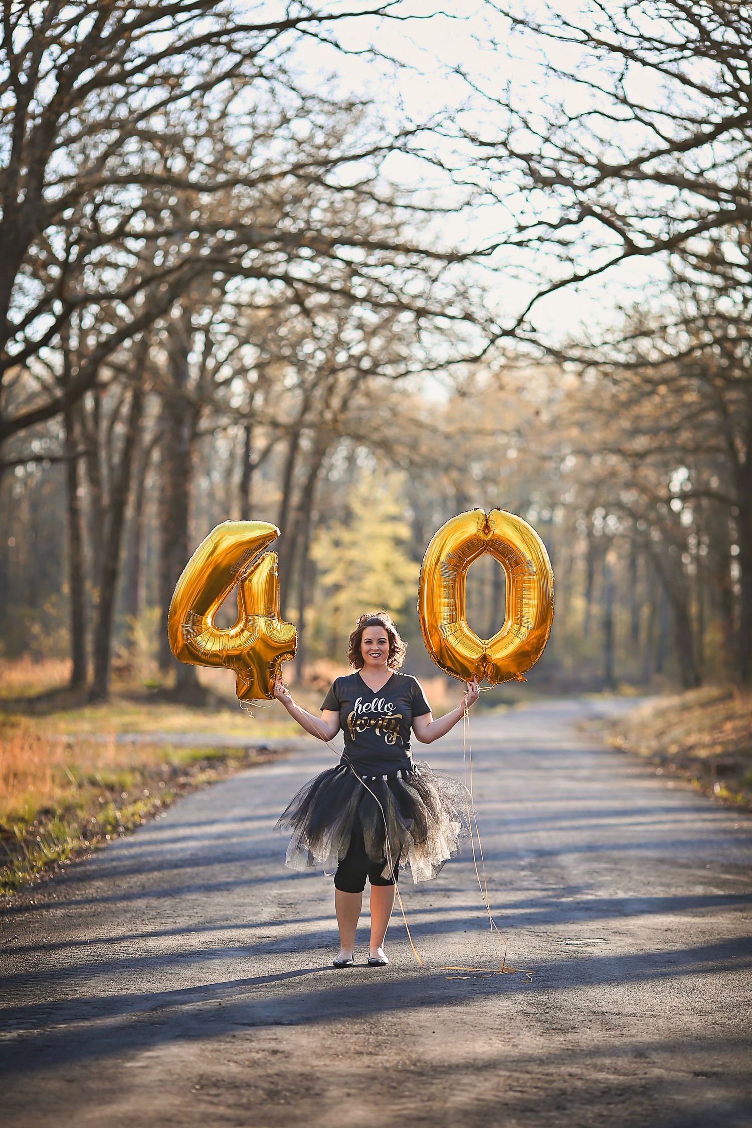 Woman celebrating 40th birthday holding gold number balloons on a tree-lined road wearing a tutu skirt.