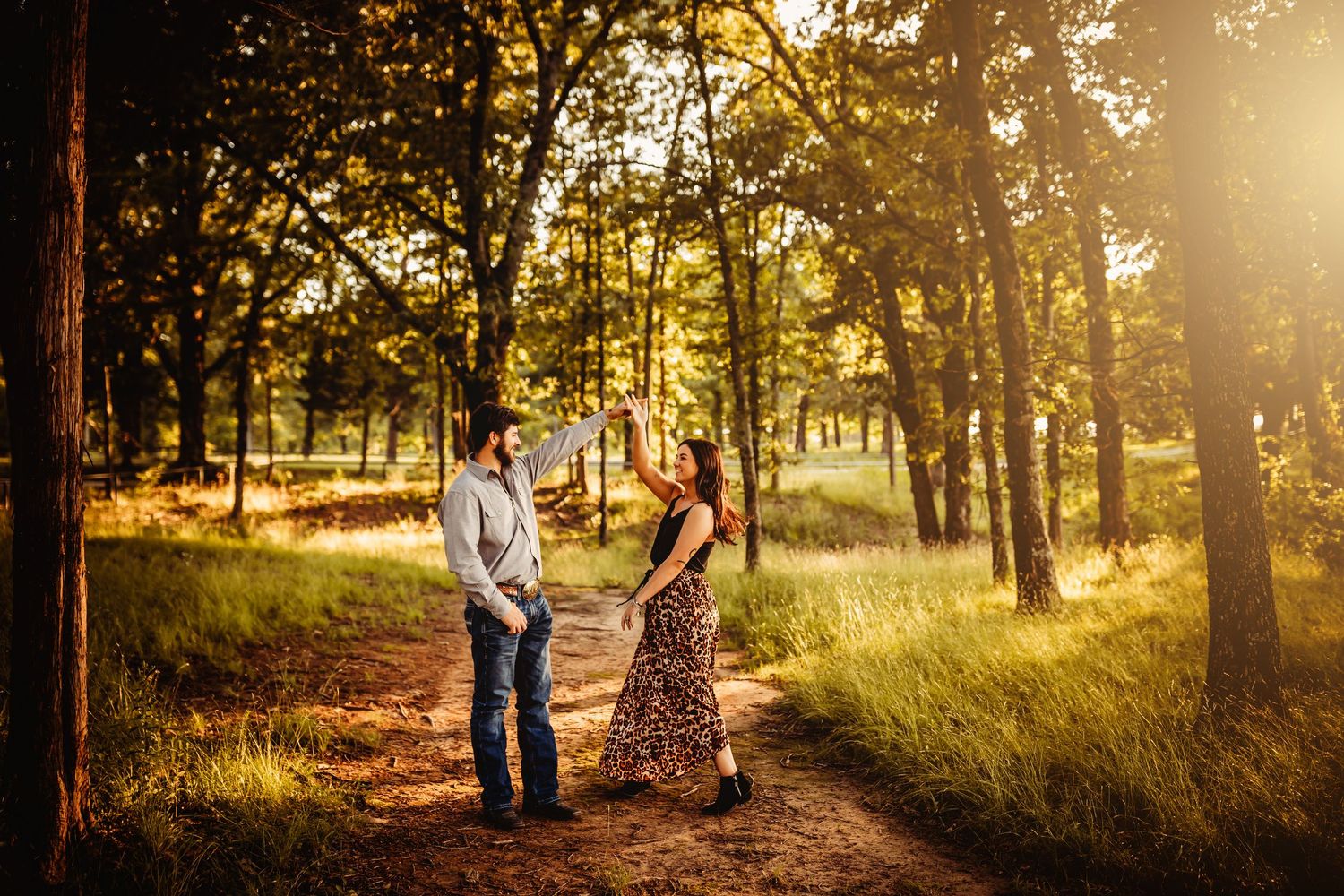 Couple standing on a sunlit forest path surrounded by tall trees during a golden hour outdoor engagement session.