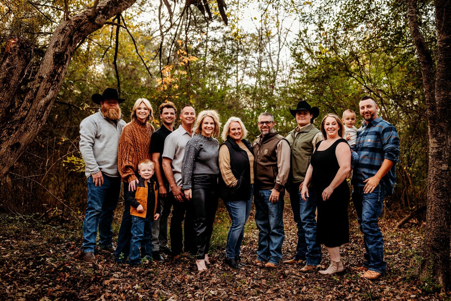 Large family group posing together outdoors in autumn woods, wearing casual western attire, smiling among fall foliage.