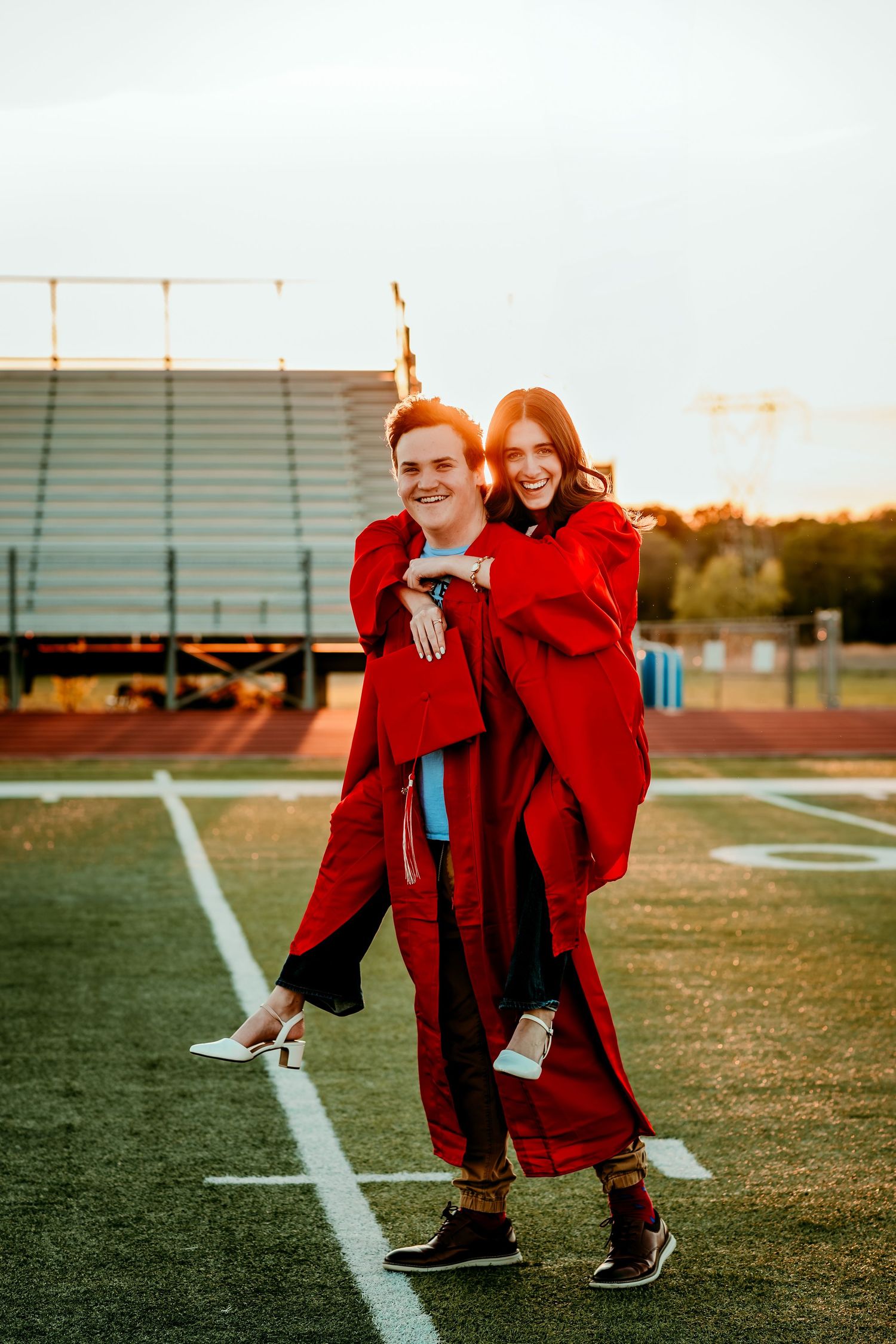 Two high school graduates in red caps and gowns celebrate on a football field at sunset, posing joyfully.