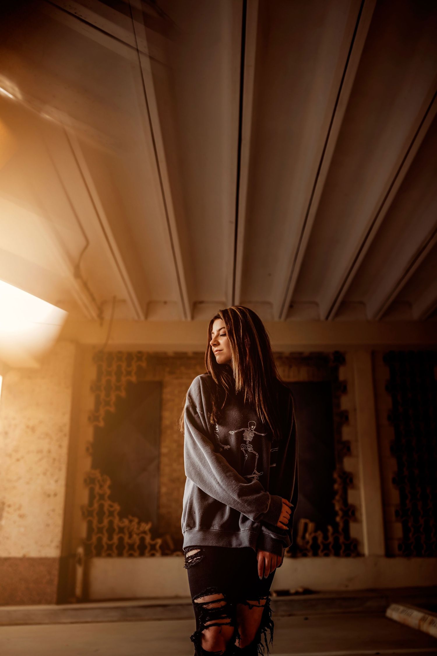 Young woman in gray hoodie and ripped jeans poses in moody urban setting with warm golden light.